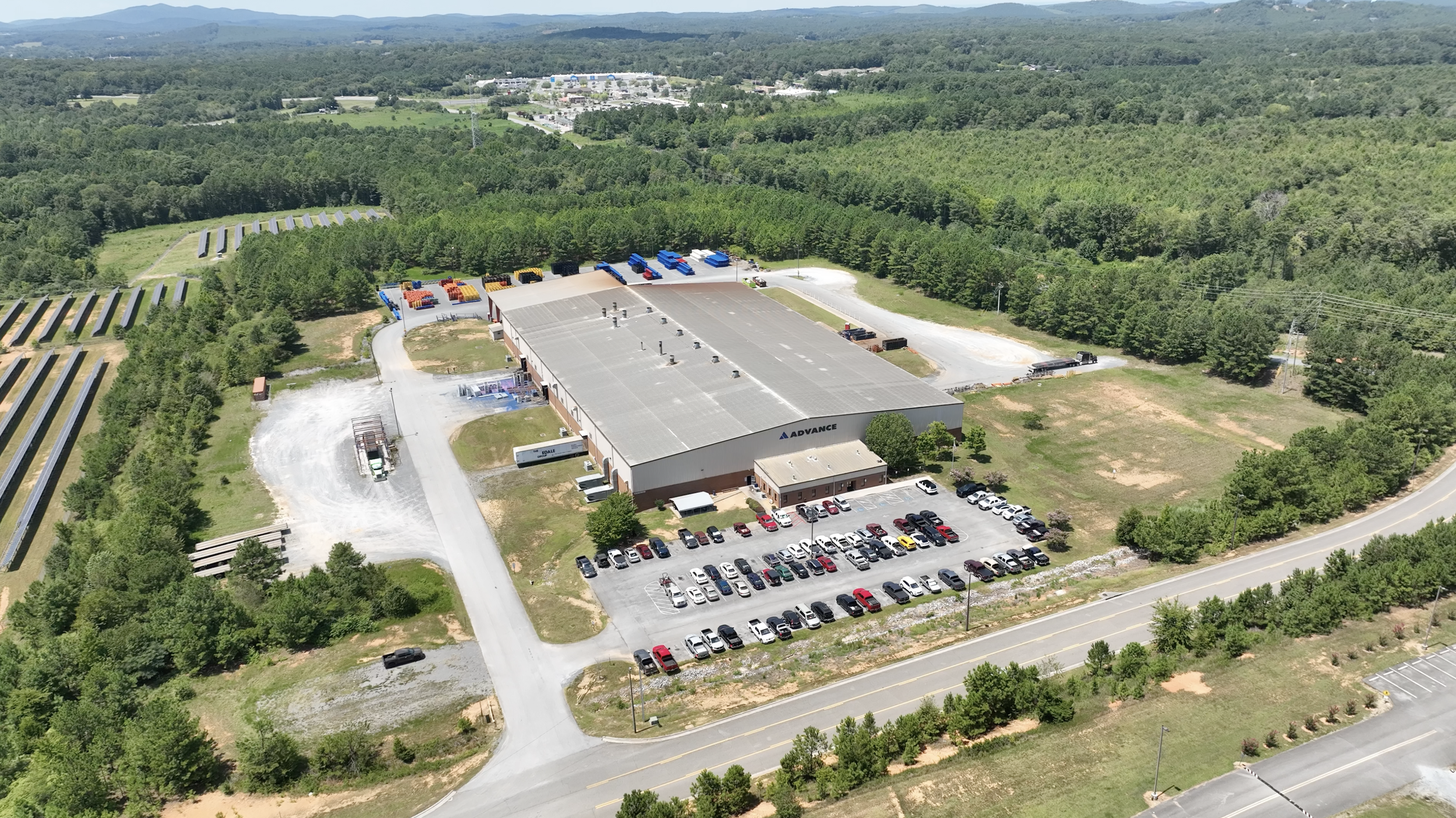 Aerial view of an industrial building with parking lot, surrounded by green trees and fields, with mountains in the background.