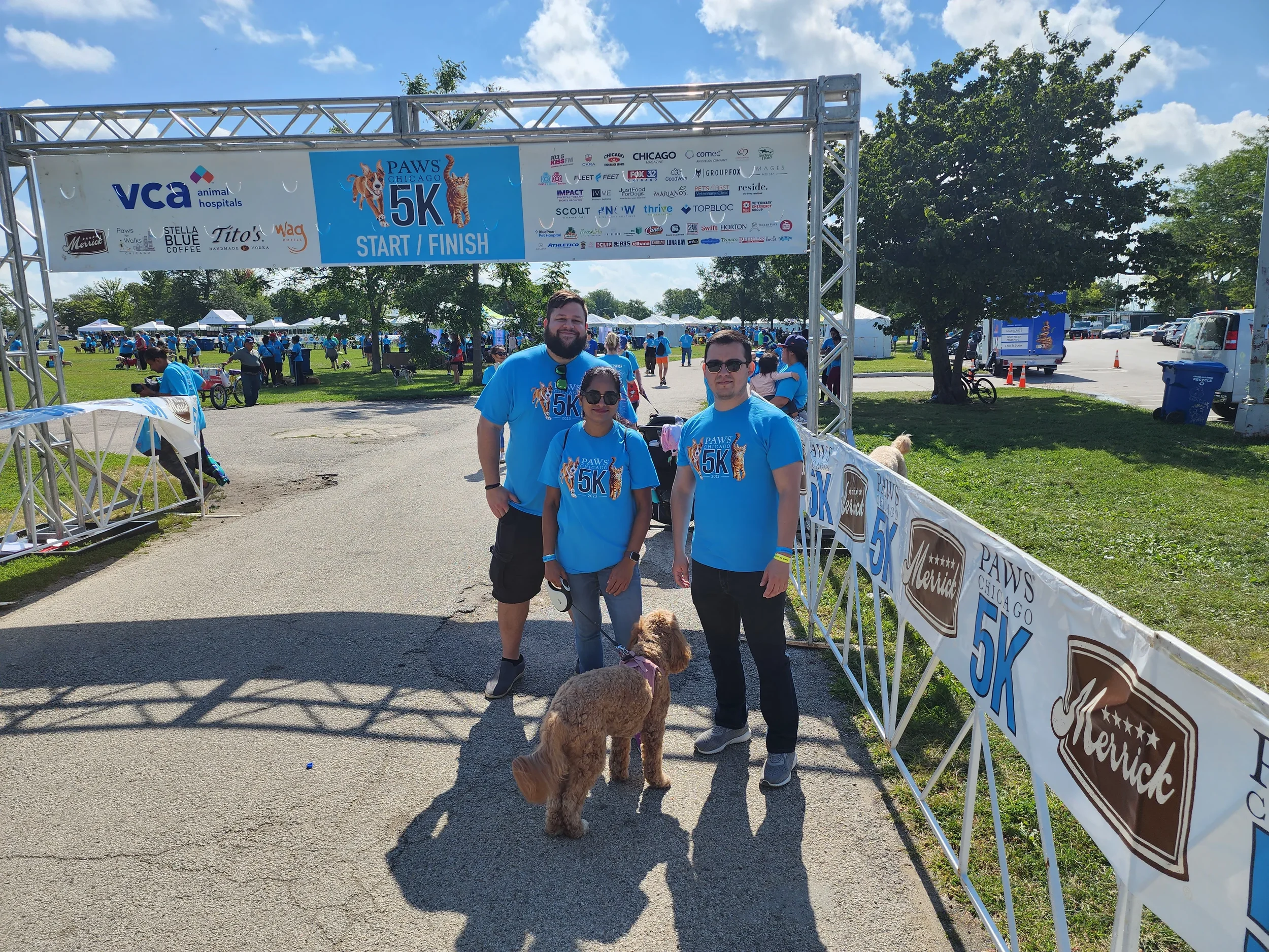 Group of people, including a man, woman, and child, standing under a park event banner for a 5K walk/run pet charity event, PAWS Chicago. They are dressed in blue T-shirts, with some holding dogs on leashes, and are smiling at the camera. There are tents and trees in the background on a sunny day.