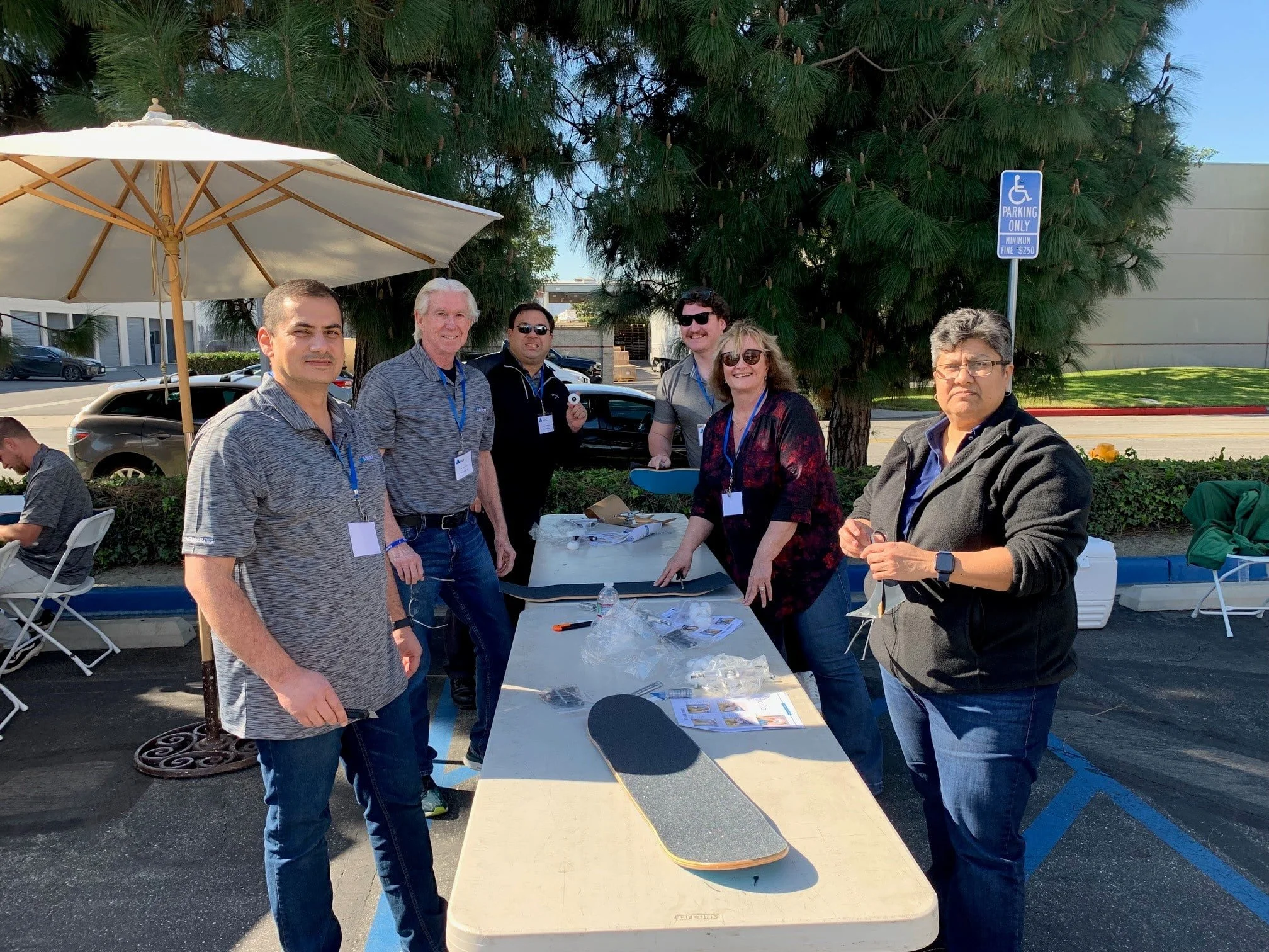 Group of six people standing around a table outdoors under a large patio umbrella, with trees and parked cars in the background.