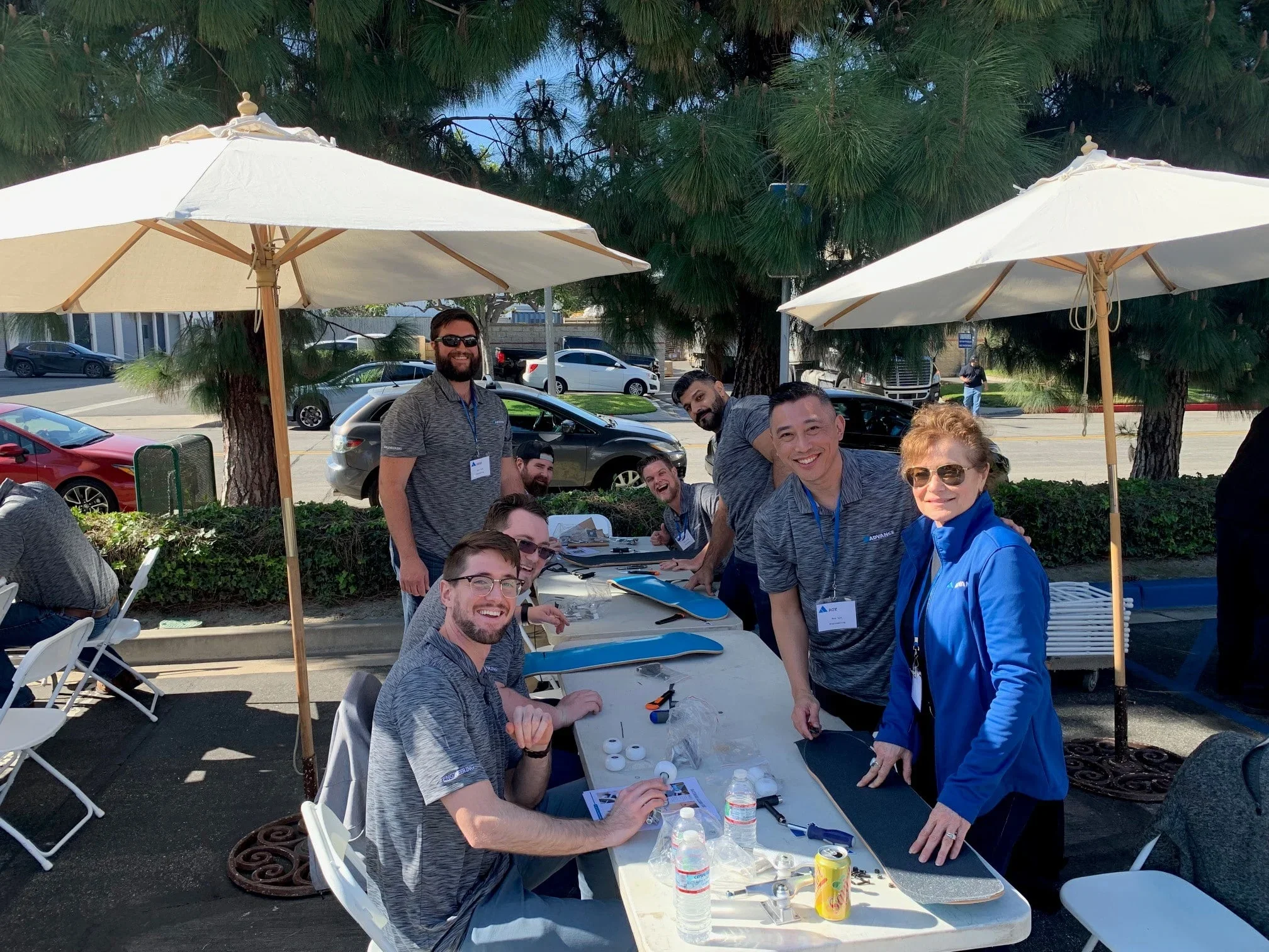 Group of people working on electronic devices and assembling objects at an outdoor table under umbrellas, with cars parked in the background and trees overhead.