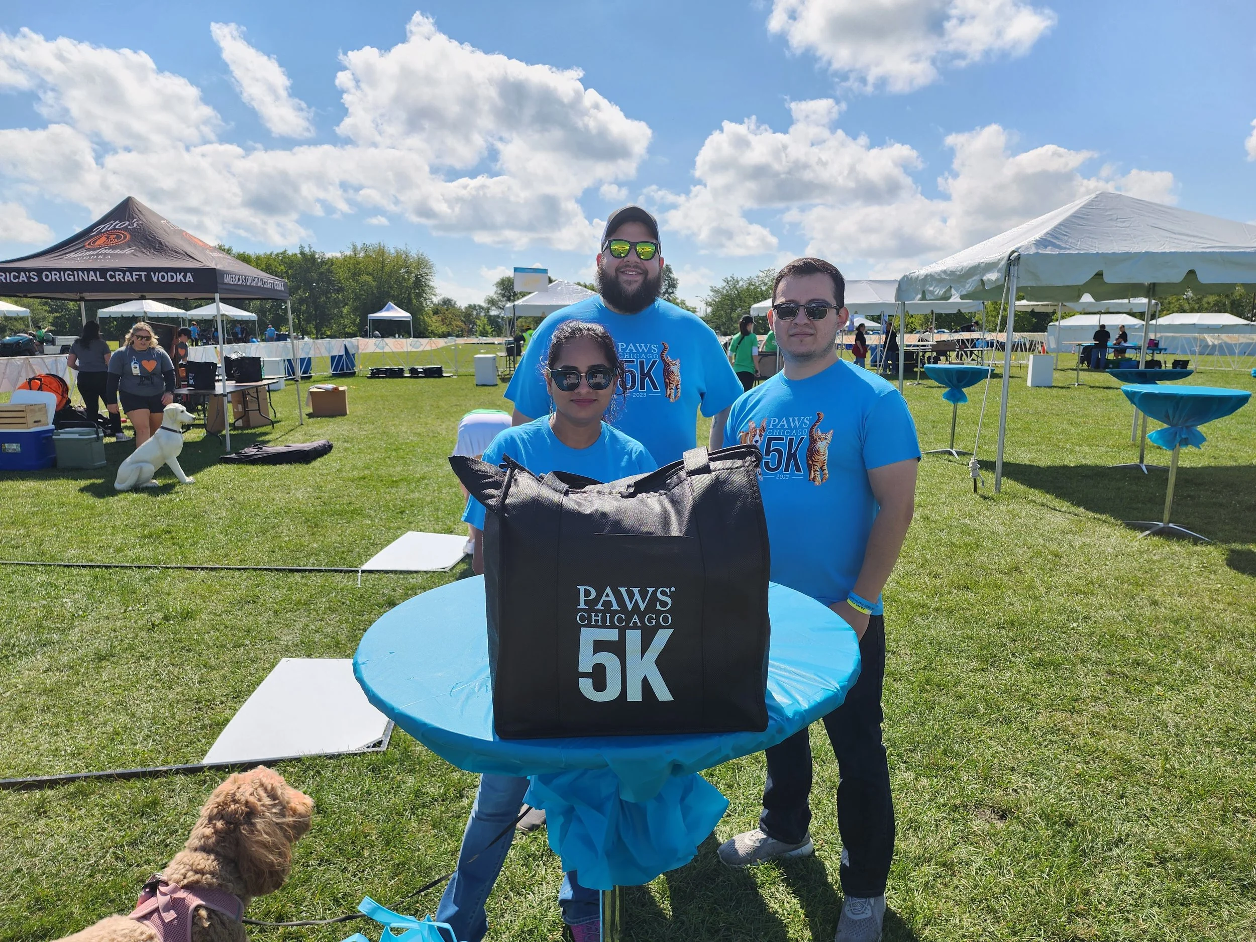 Three people standing together at an outdoor event under a blue sky with clouds. They are wearing matching blue race shirts that say 'PAWS Chicago 5K 2023'. A man with a beard, a woman, and a man all wear sunglasses and smile at the camera. There are tents and people in the background, and a small dog with a brown coat is sitting on the grass in front of them.