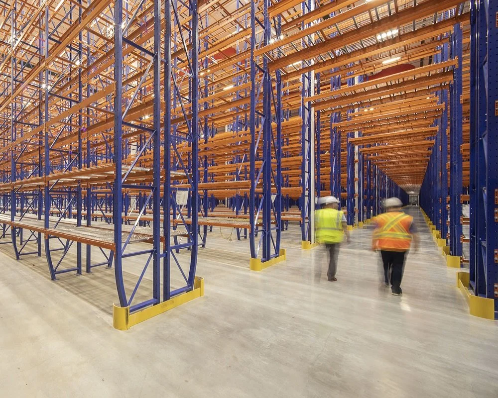 View of an empty warehouse with blue and orange metal shelving units and two workers wearing yellow safety vests and white helmets walking down a pick tunnel.