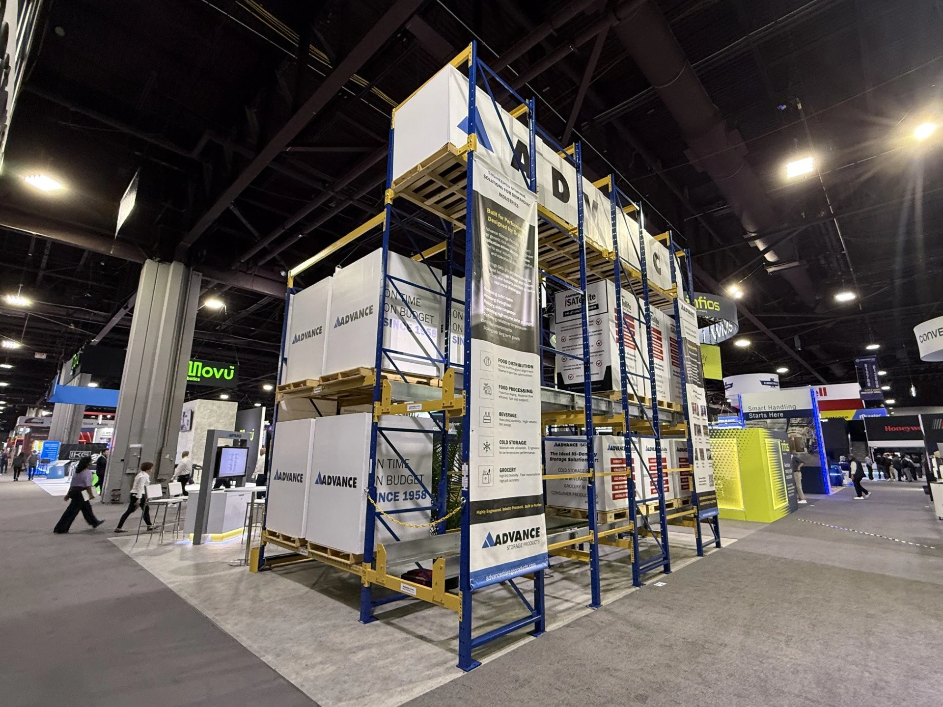 Indoor exhibition with a metal shelving unit displaying banners for 'Advance Storage Products' and other booths in the background, with visitors walking through the space.