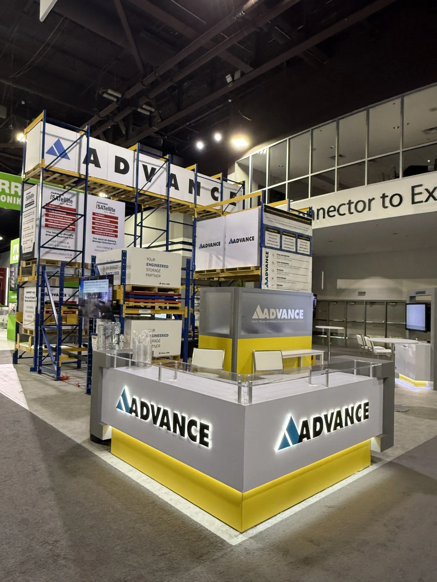 Trade show booth with signage for Advance, featuring a white and yellow display counter and scaffolding for signage, with part of a large sign reading 'Connector to Ex...' in the background.