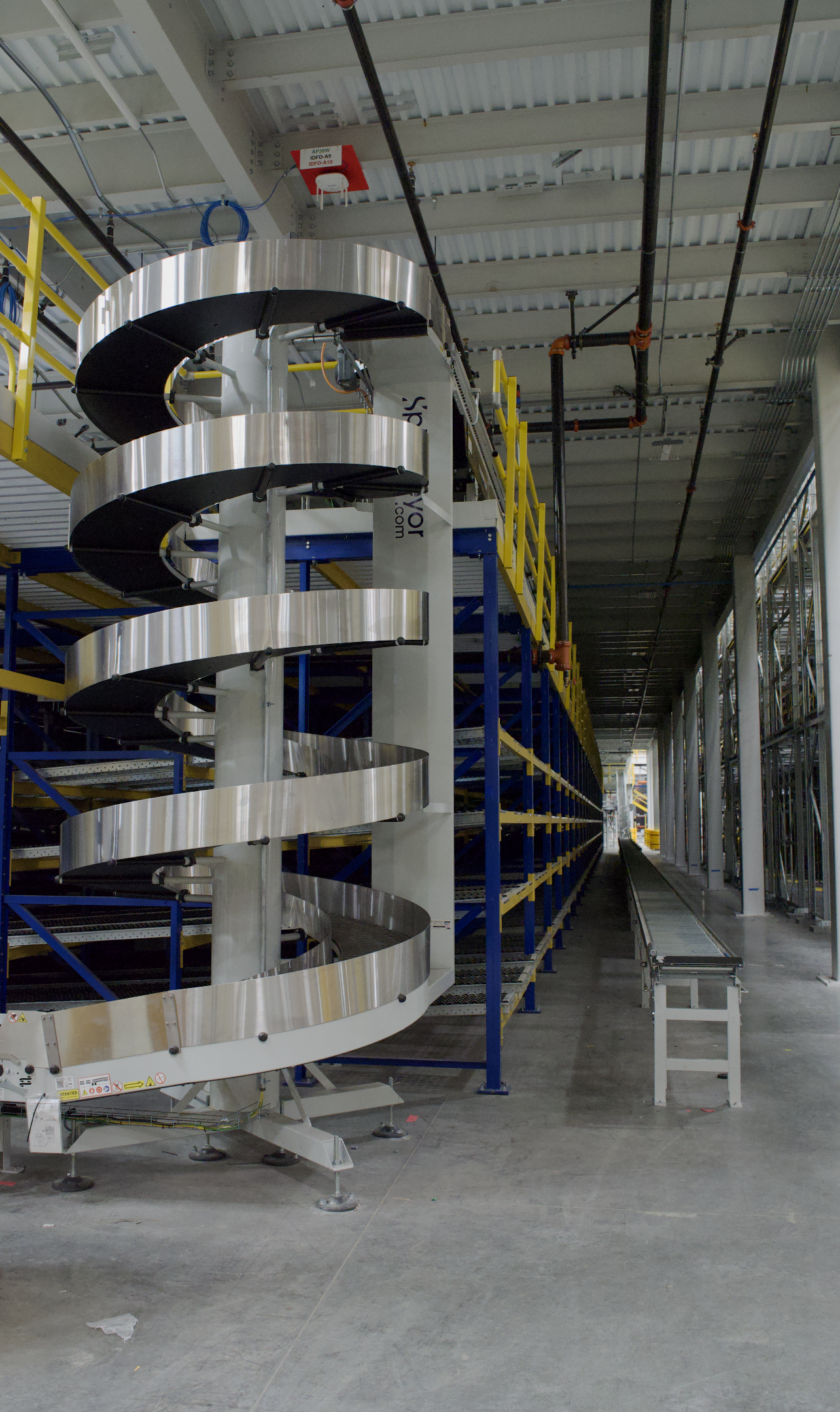 Industrial warehouse with a metallic spiral conveyor or slide, adjacent to blue and yellow shelving units, and a metal roller conveyor on the right side, with visible pipes and wiring on the ceiling.