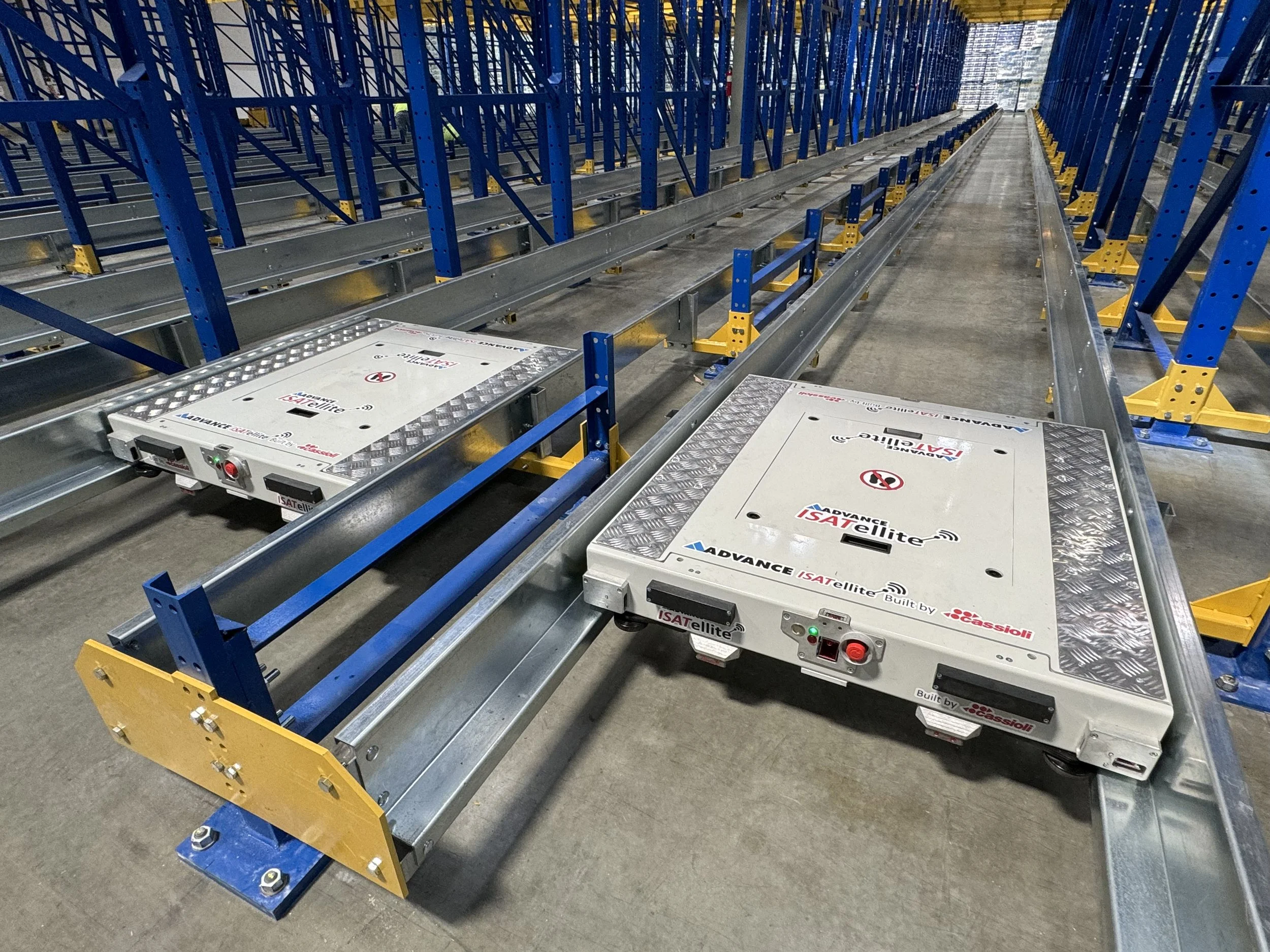 Industrial warehouse with blue metal racks and automated satellite delivery systems on a concrete floor.