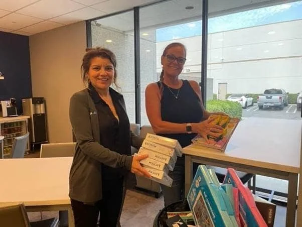 Two women standing inside a library or bookstore near a large window, holding and organizing books, with bookshelves and a parking lot visible outside.