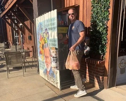 A man wearing sunglasses, a dark t-shirt, gray pants, and white sneakers standing outside a shop, holding a plastic bag in his right hand. The shop has a colorful painted mural on the side and a red brick exterior.