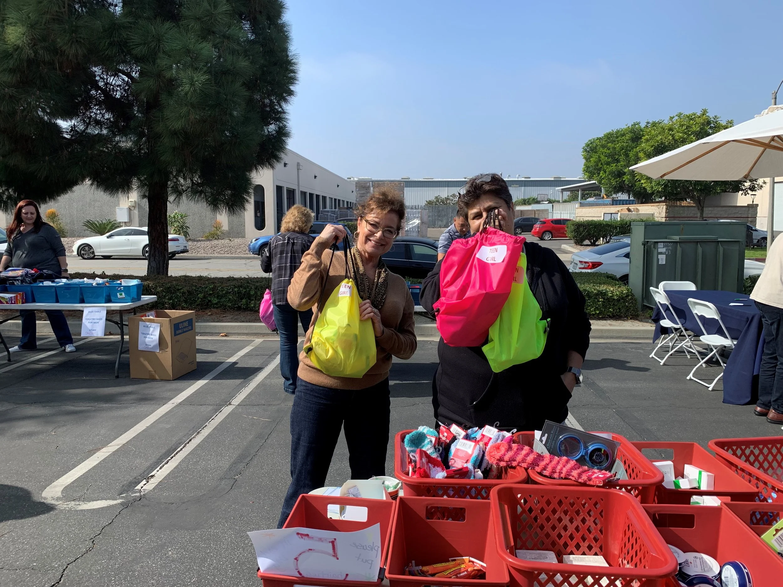 Two women standing behind red bins filled with socks and other items at an outdoor event, holding bright pink and yellow bags labeled 'Teen Girl.' There are other people in the background, and shade can be seen on the right side of the image.
