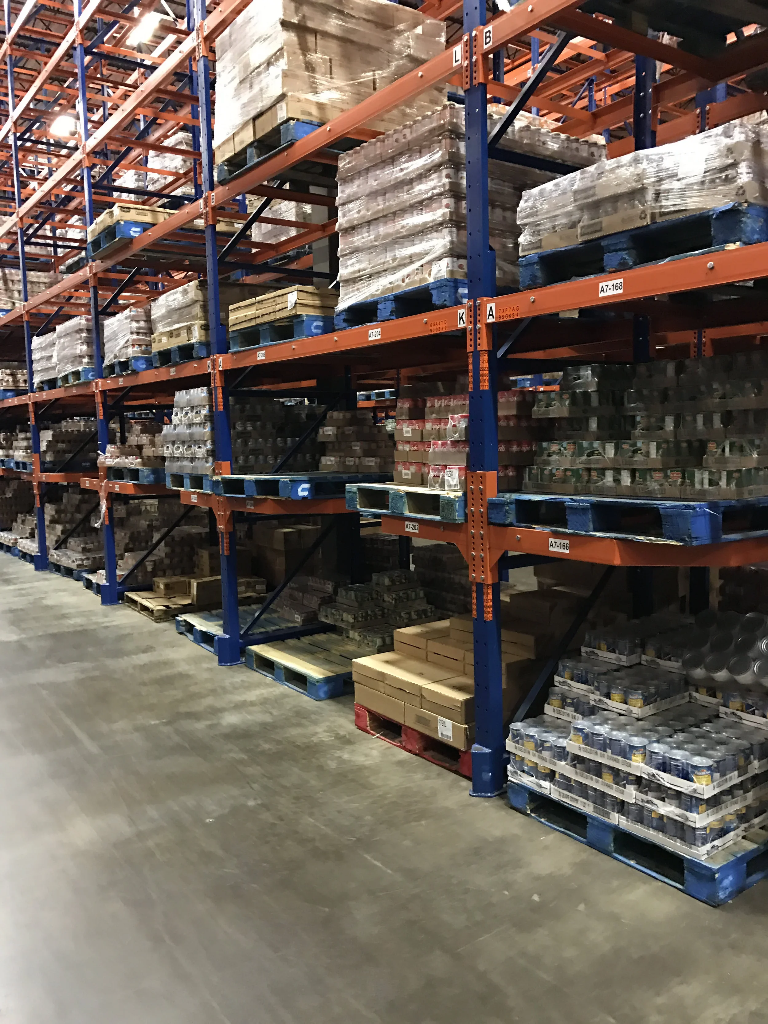Warehouse aisle with tall shelving units stocked with pallets of various items wrapped in plastic, including boxes and metal objects.