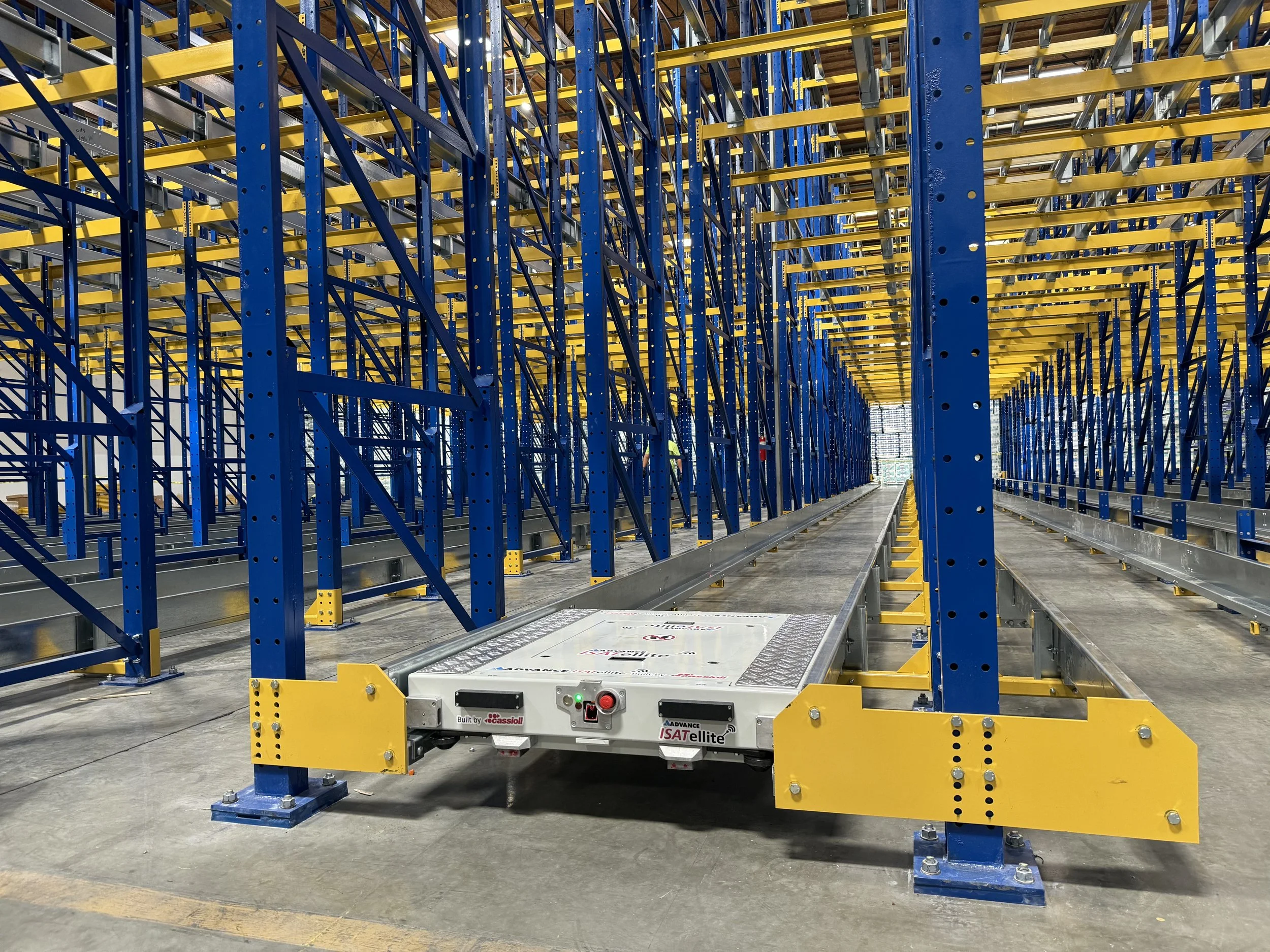 Interior view of a large warehouse with blue and yellow metal shelving racks and a small autonomous robotic vehicle in the center.
