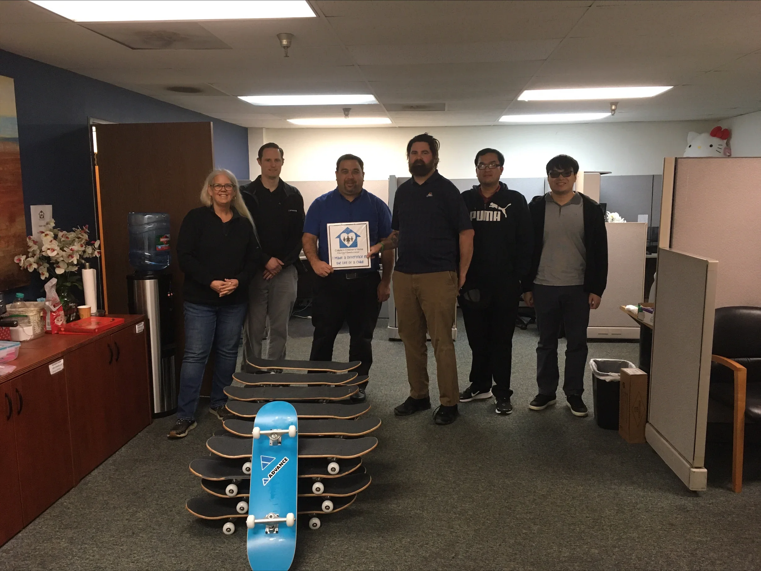 Group of six people standing in an office, holding a sign that reads 'Made a difference in the Life of a Child,' with skateboards placed in front of them. The background has cubicles, a water dispenser, and Hello Kitty decorations.
