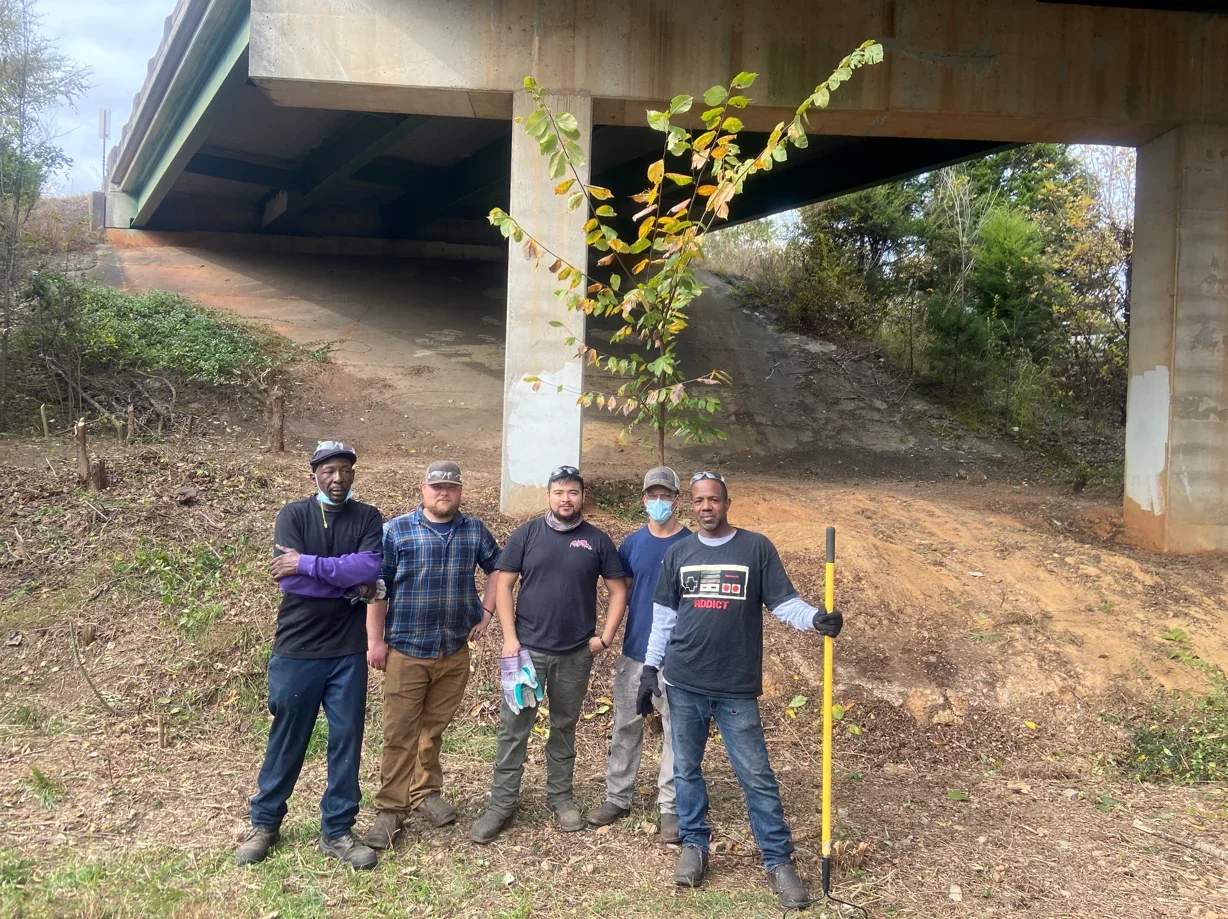 Five men standing outdoors under a bridge, some holding tools, in a dirt area with small trees.