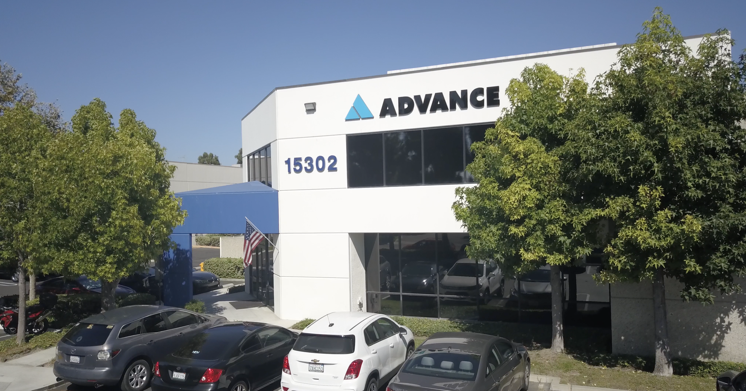 Photo of an office building with the name 'Advance' on it. The building has large windows, white walls, and a blue accent. There are trees in front and a parking lot with several parked cars and an American flag near the entrance.