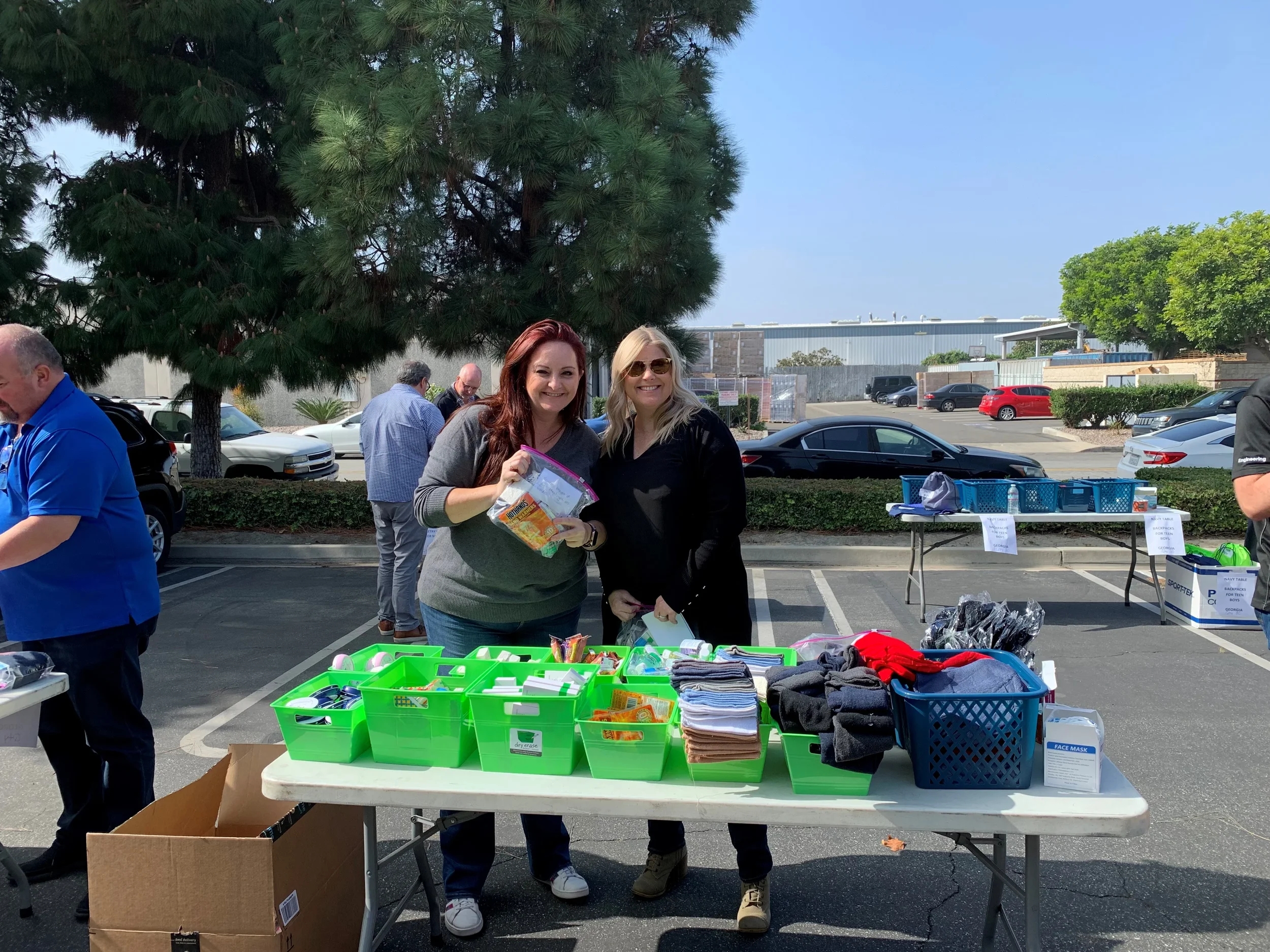Two women stand behind a table of assorted items at an outdoor sale or community event, smiling at the camera. The table has containers filled with small objects, socks, and other items. There are other people in the background and a parking lot with cars, trees, and a building nearby.