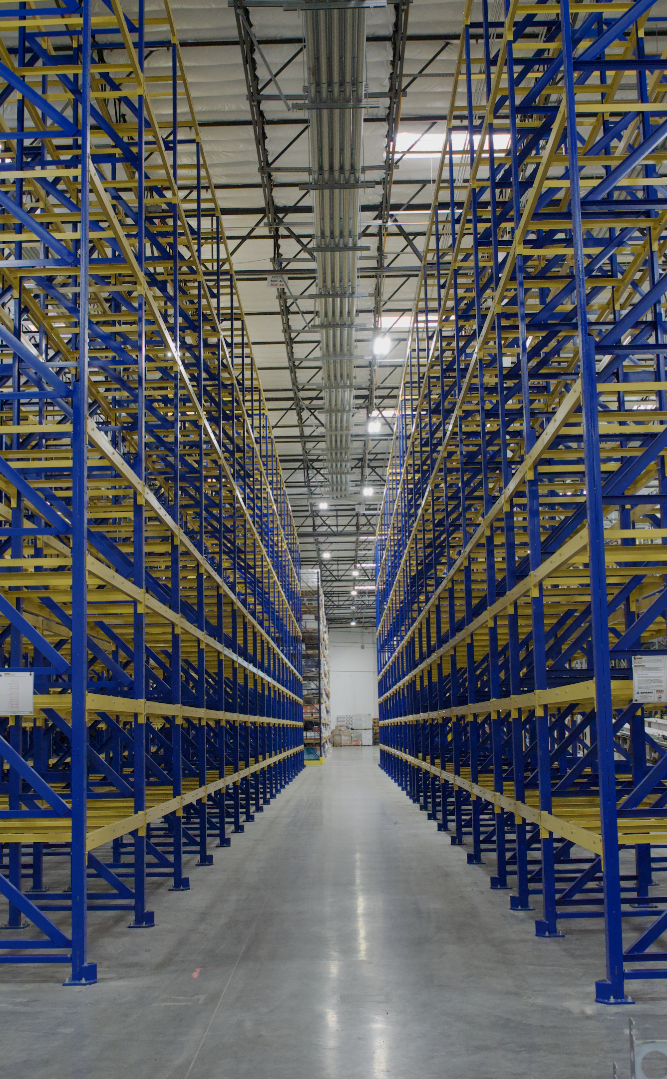 Long aisle in a warehouse with tall blue and yellow metal shelving units on both sides, and a high ceiling with industrial lighting.
