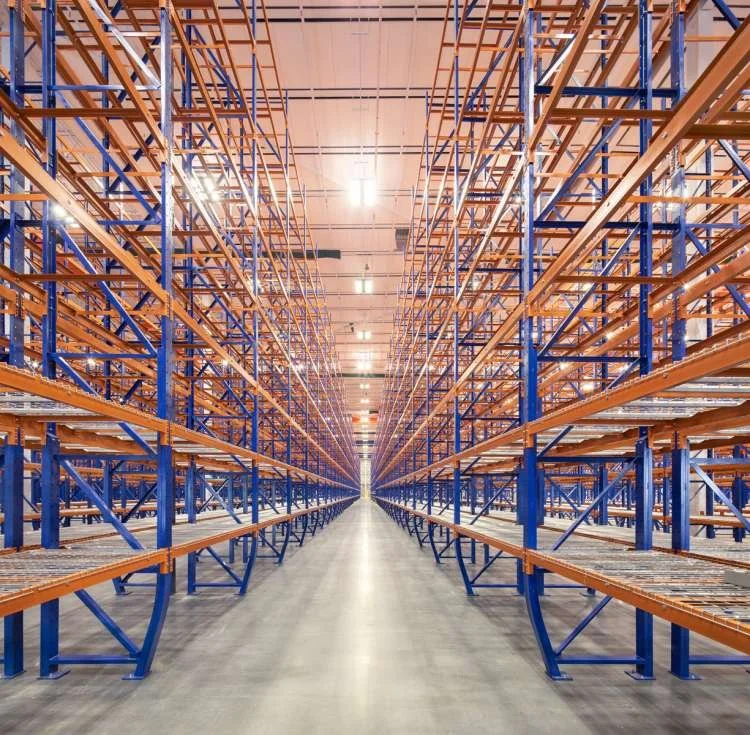 A large, empty warehouse with tall blue and orange metal shelving units arranged in parallel rows, extending into the distance.