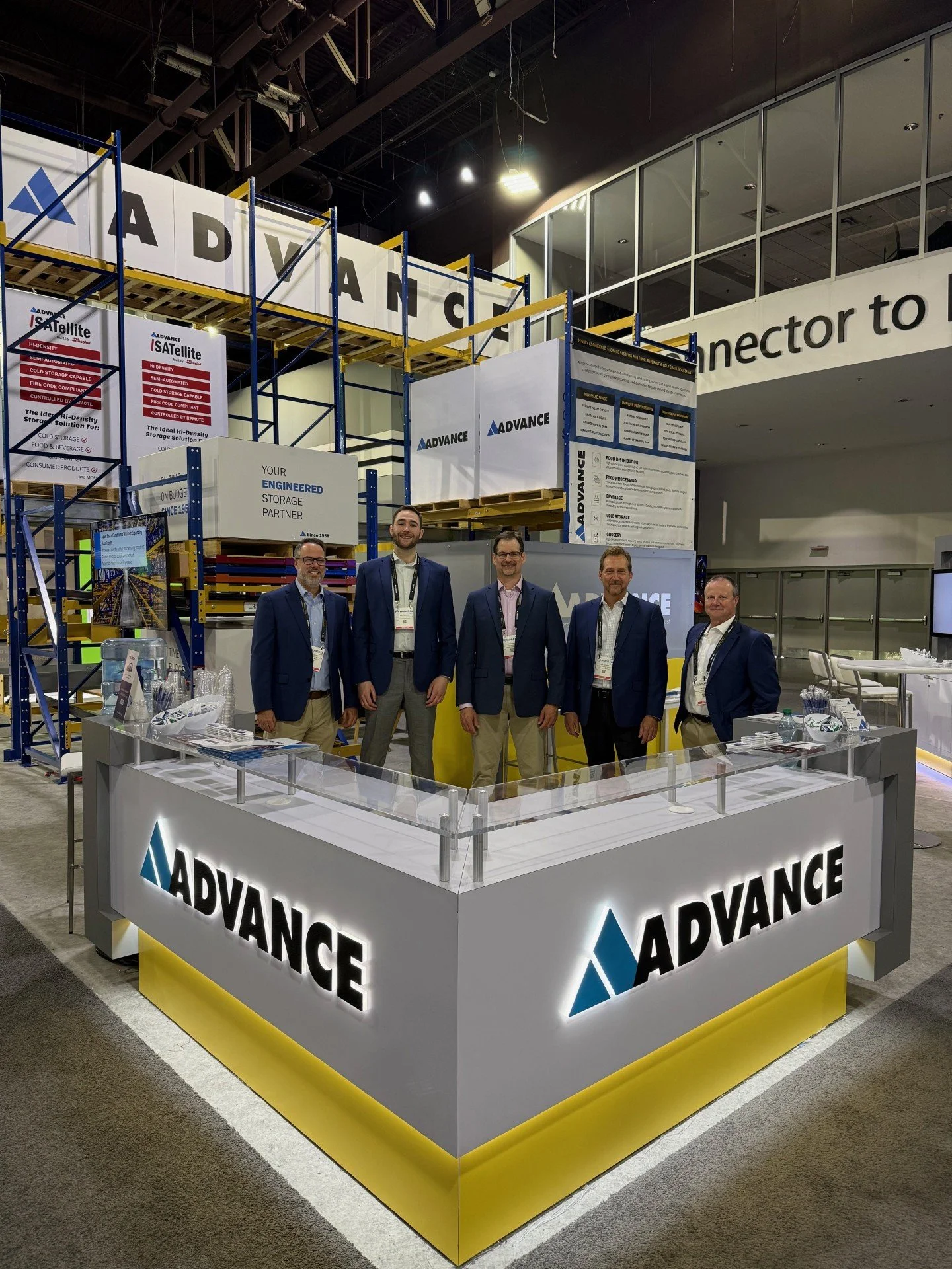 Trade show booth for Advance with four men in business attire standing behind a white counter with the company logo, surrounded by shelving units and promotional displays.