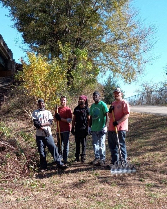 Five people outdoors in a sunny area with trees, holding gardening tools and standing on dirt and grass.