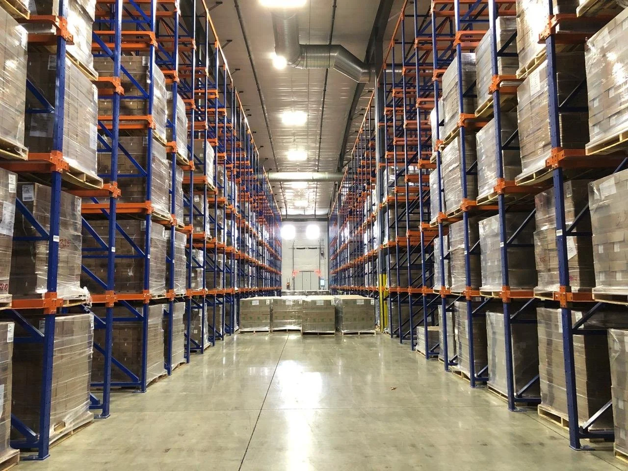 A large warehouse with tall metal shelving units stacked with pallets of goods, in drive-in racking.