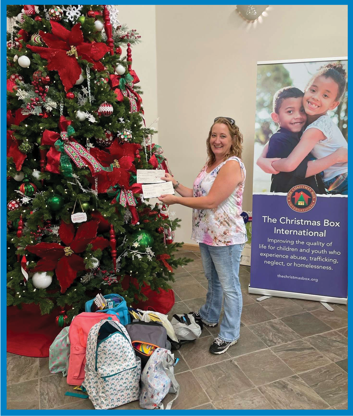 A woman standing next to a decorated Christmas tree holding two donation checks, with backpacks and school supplies on the floor. A banner for The Christmas Box International is visible in the background.