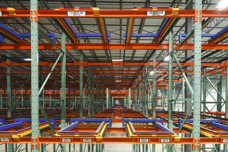 Empty industrial warehouse shelving units, organized in rows, with orange and purple metal pushback racks.