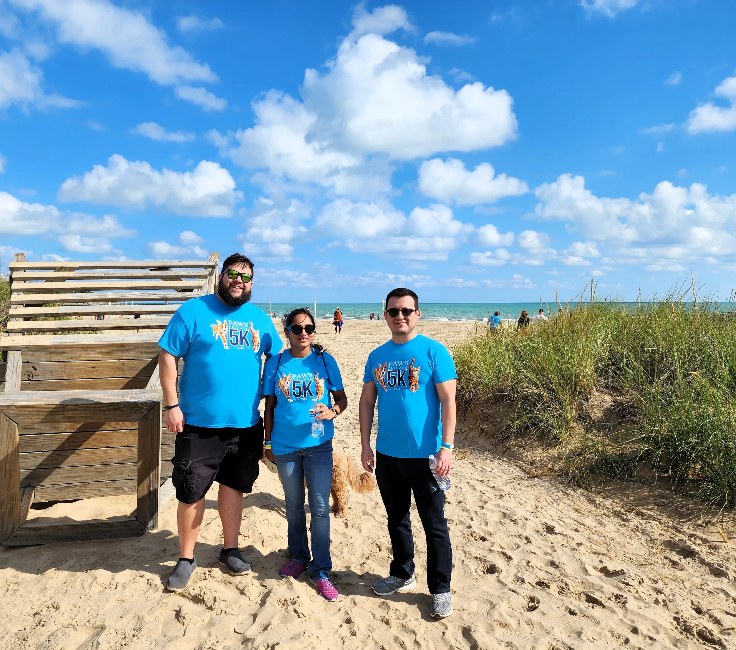 Group of four people standing on a sandy beach near a wooden structure, with a dog, under a bright blue sky with scattered clouds.