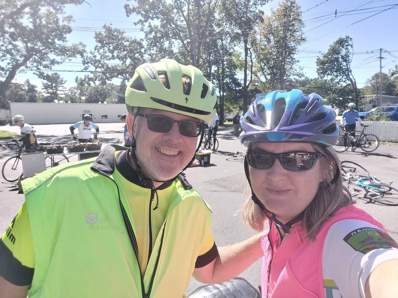 Two cyclists taking a selfie, both wearing helmets and sunglasses, with bicycles and other cyclists in the background on a sunny day.