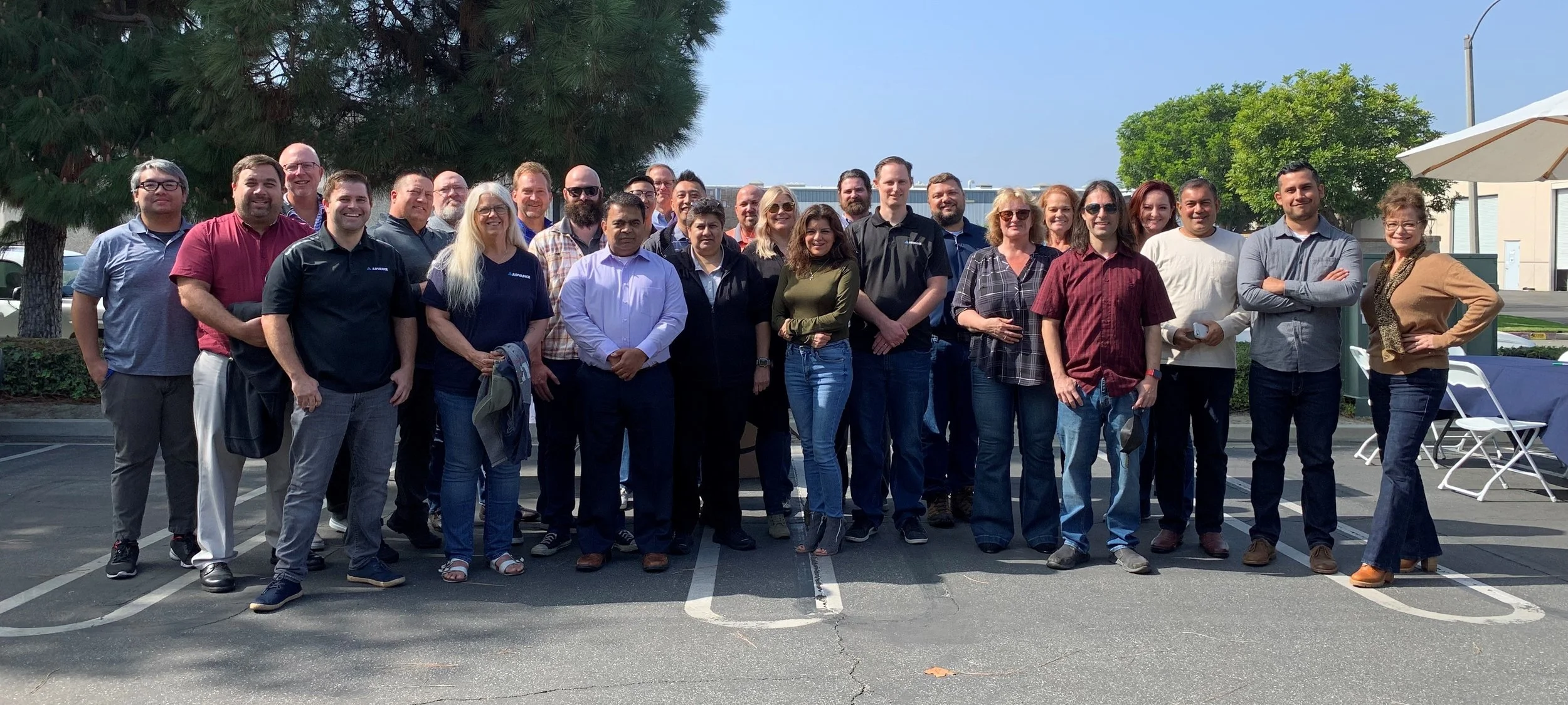 A group of about 20 diverse people standing outdoors in a parking lot during daytime, smiling for the photo, with trees and a building in the background, some wearing company-branded shirts.