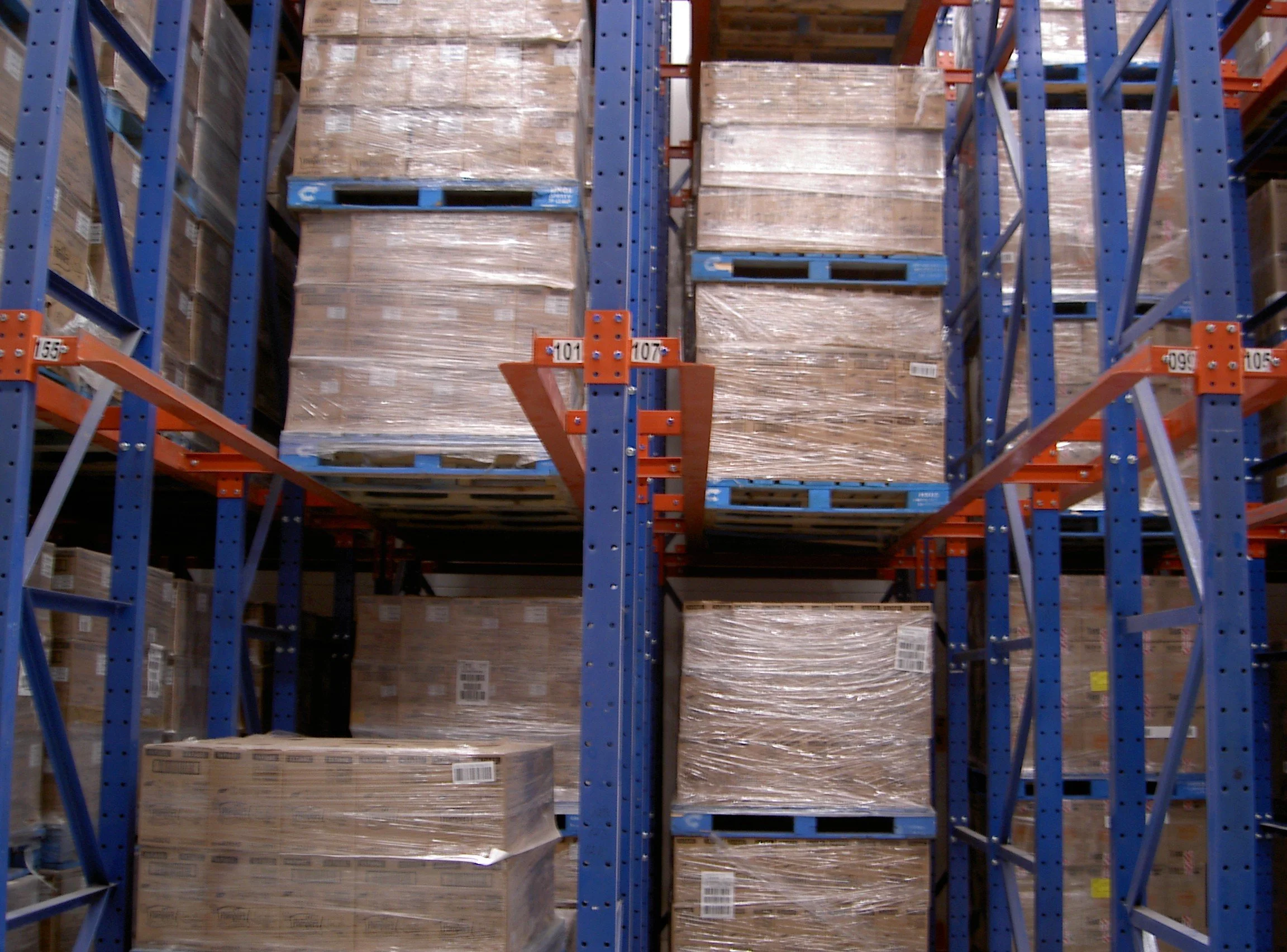 Warehouse storage shelves filled with stacked boxes wrapped in plastic, layered within drive-in racking.