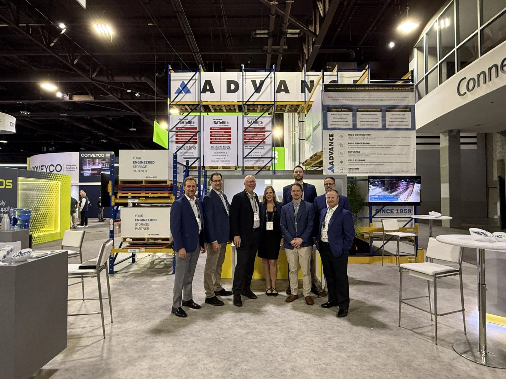 Group of seven professionals standing together at a trade show or conference, smiling for the photo, with exhibition booths and banners in the background.