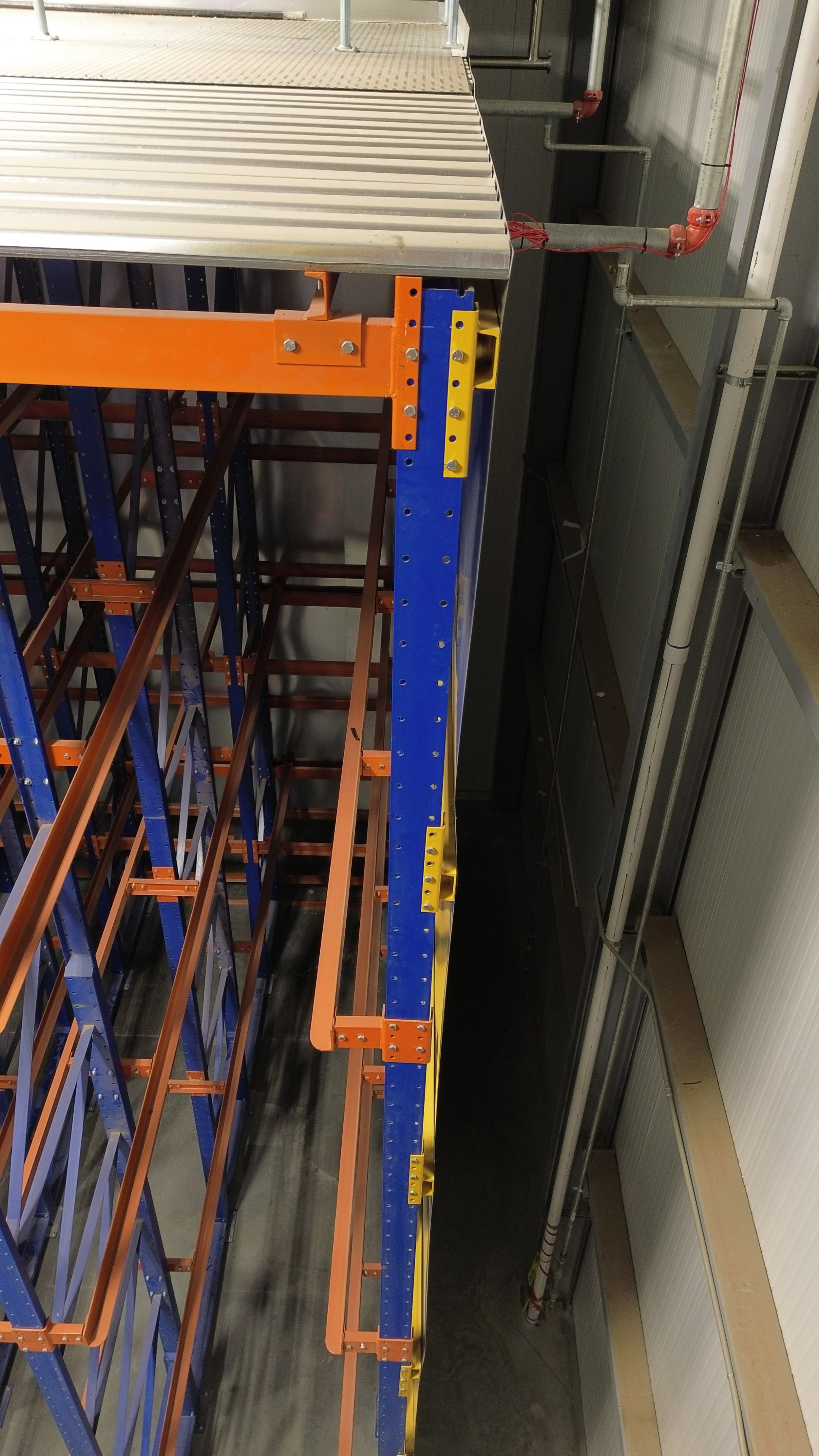 View of industrial storage racks with orange, blue, and yellow metal framing in a warehouse or manufacturing facility. The racks are empty.