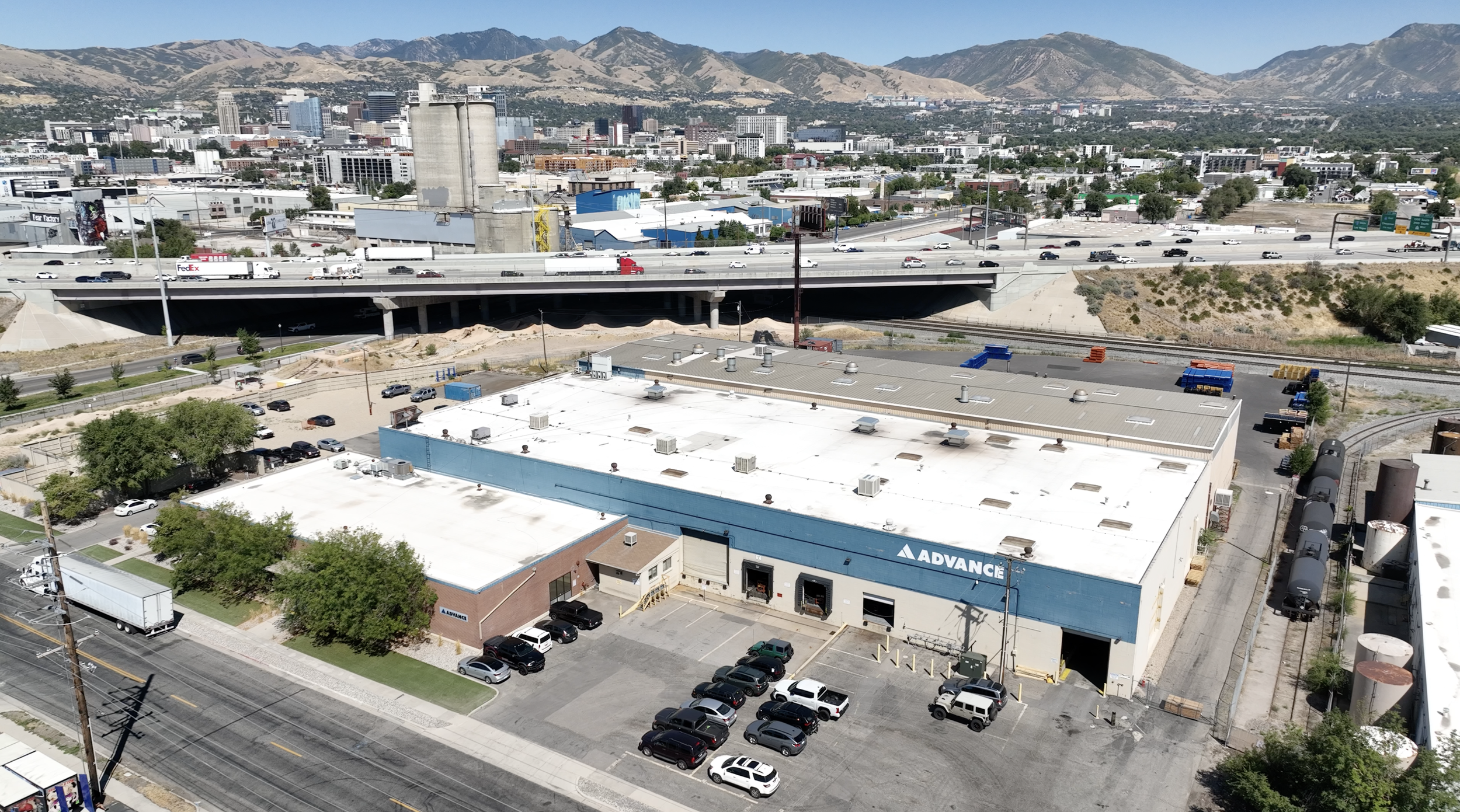 Cityscape with mountains in the background, a highway with moving cars, and an industrial building with a blue and white sign reading 'ADVANCE' in the foreground.