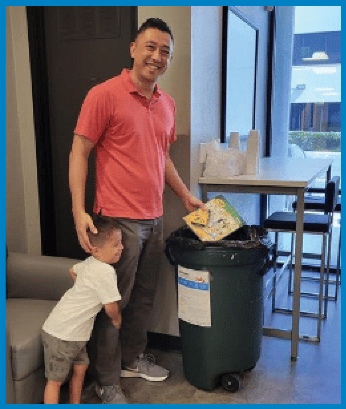 A man in a red polo shirt and a young boy with a white shirt and shorts, standing next to a green trash can in a room with a table and chairs near a window. The man is holding a children's book and smiling.
