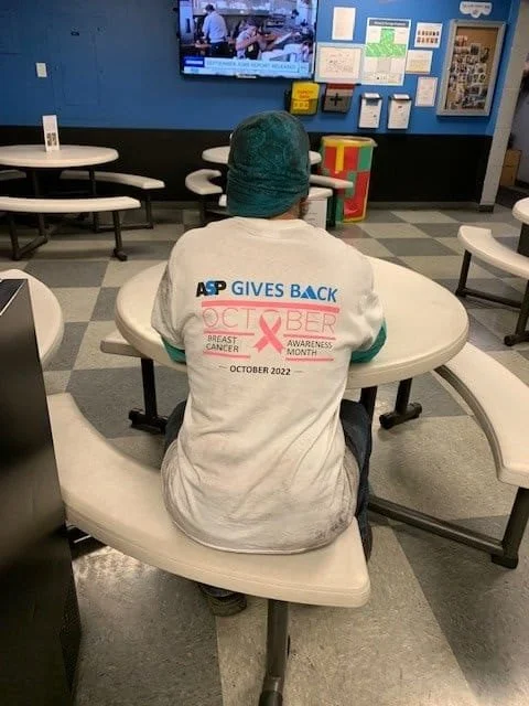 Person sitting at a table in a waiting room, wearing a white shirt and a teal surgical cap, with a breast cancer awareness logo and text on the back for October 2022