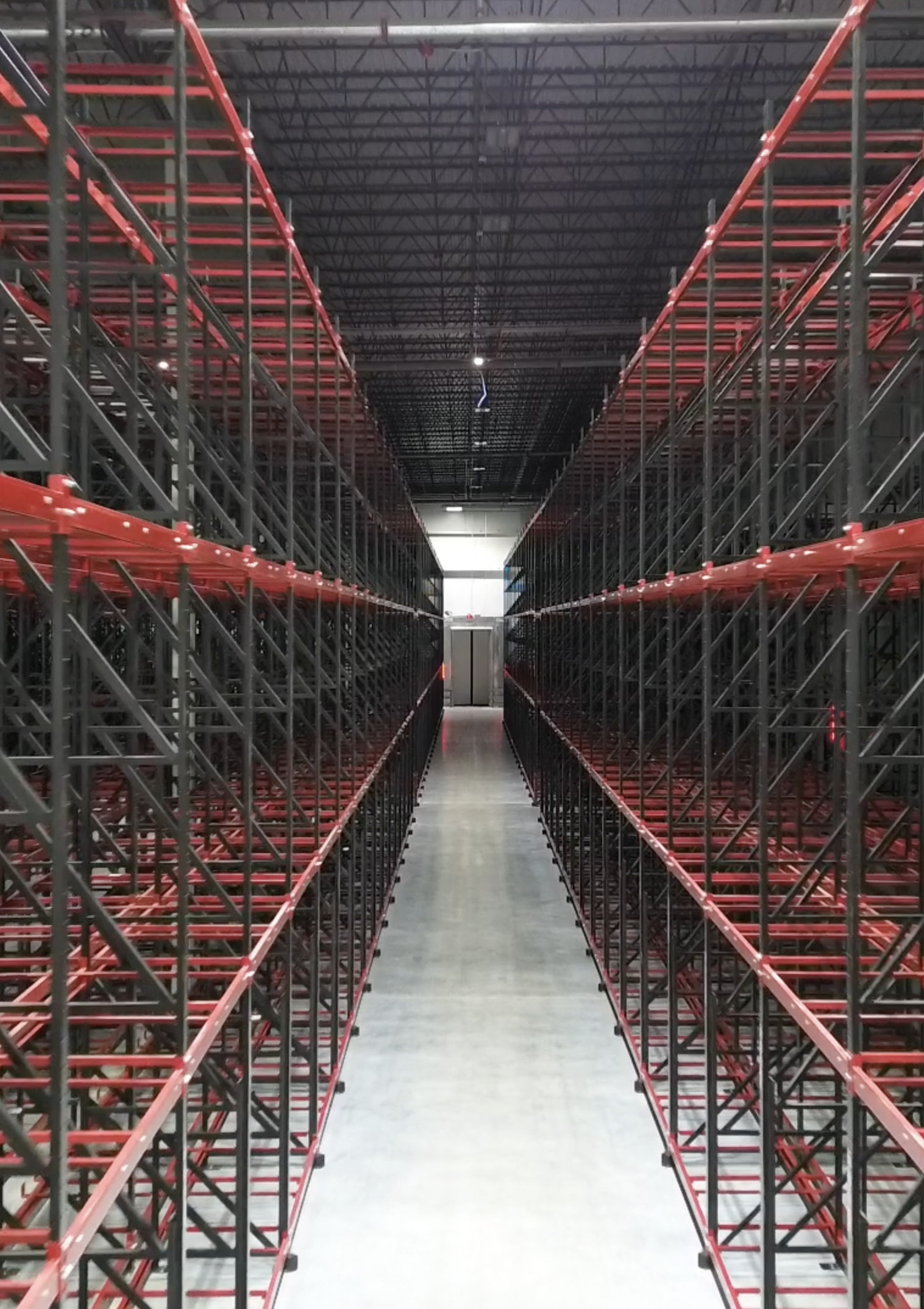 Empty warehouse with multiple industrial steel shelving units on both sides of a central aisle, painted with red accents, under a high metal roof with visible support beams.