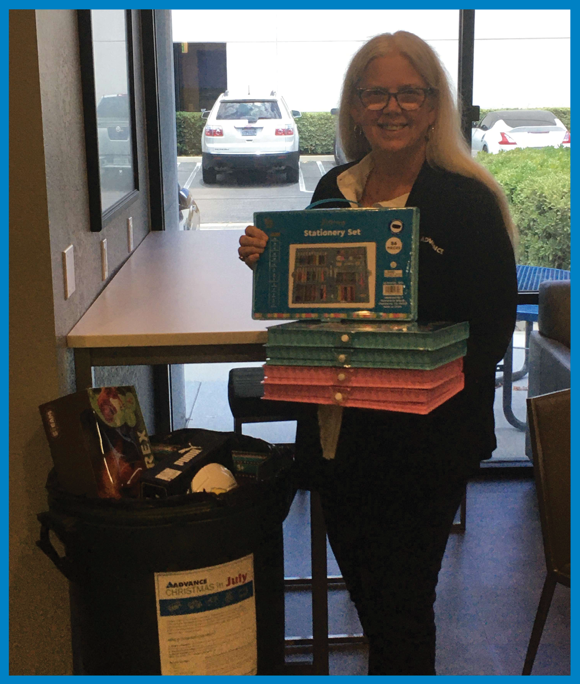 A woman with long gray hair and glasses, smiling, holding a box of a children's book or toy set, standing in front of a window in a store or café. She is surrounded by stacks of similar boxed items, with a black bag containing snacks and a brochure on the table nearby. Outside the window, parked cars and a hedge are visible.