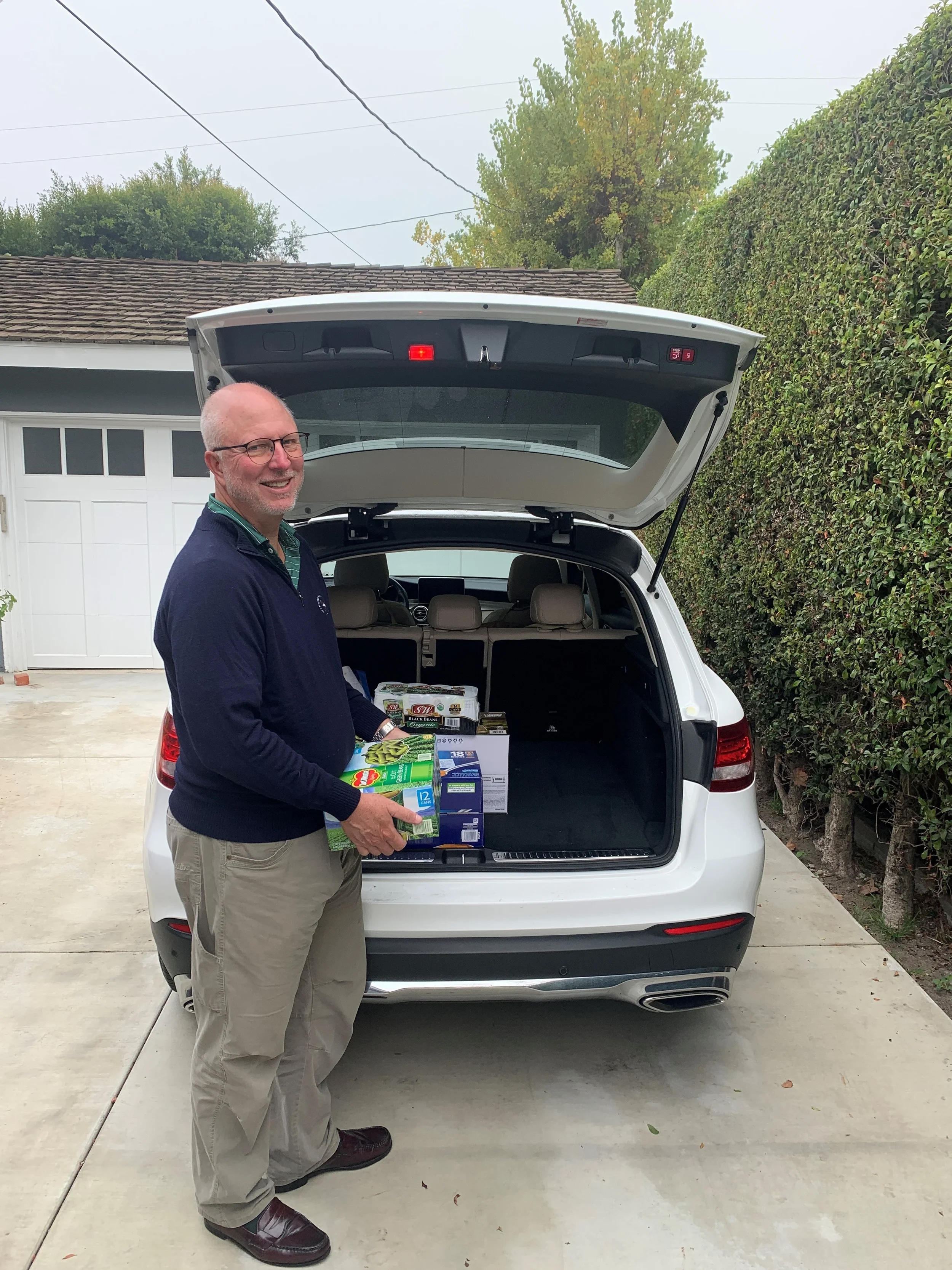 A man with glasses and a beard smiling and holding grocery items stands behind an open trunk of a white SUV parked on a driveway. The trunk contains grocery boxes and packages. The background shows a garage, a hedge, and trees.