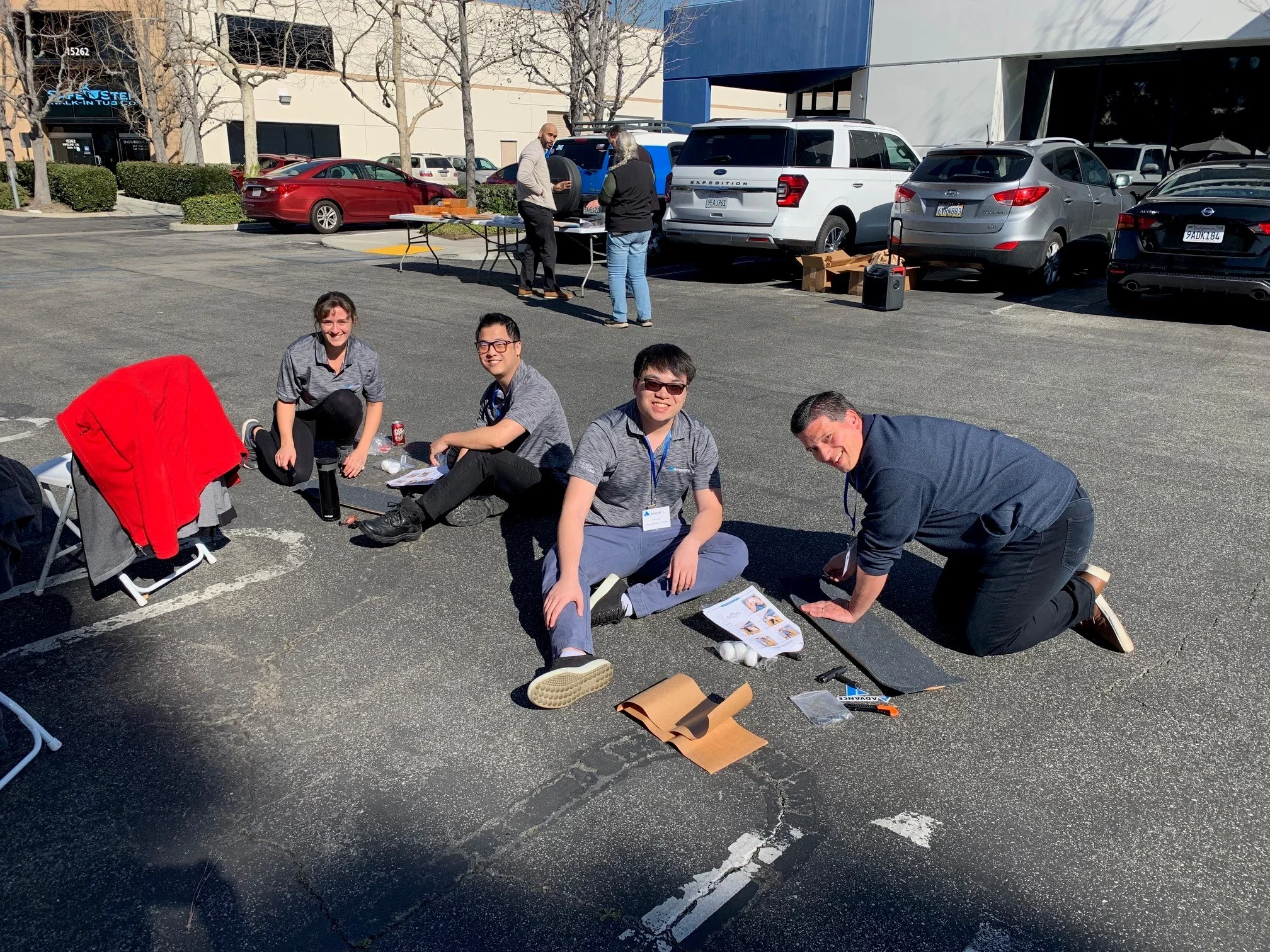 Four people sitting and lying on the ground of a parking lot, with two more people standing by tables at the background. Several parked cars are visible behind them, along with leafless trees and a building. The people appear to be participating in an outdoor activity or event, with some materials and a brochure or flyer on the ground.