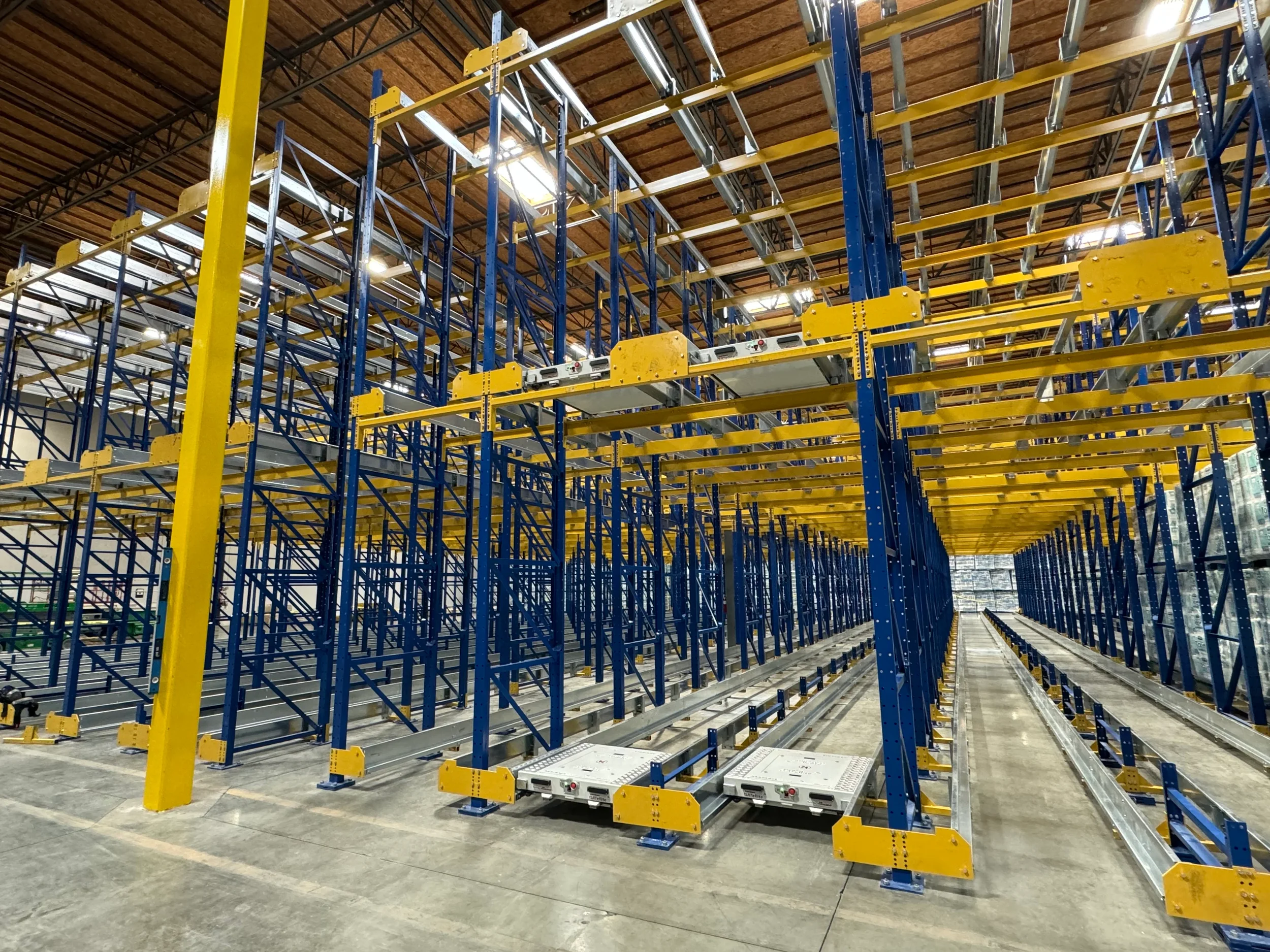 Interior of a large warehouse with metal shelving units in blue and yellow, some parts assembled and others still being constructed, under a wooden ceiling with skylights.