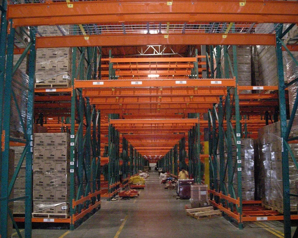 A large warehouse pick tunnel aisle through tall green and orange industrial shelving filled with boxes and pallets.