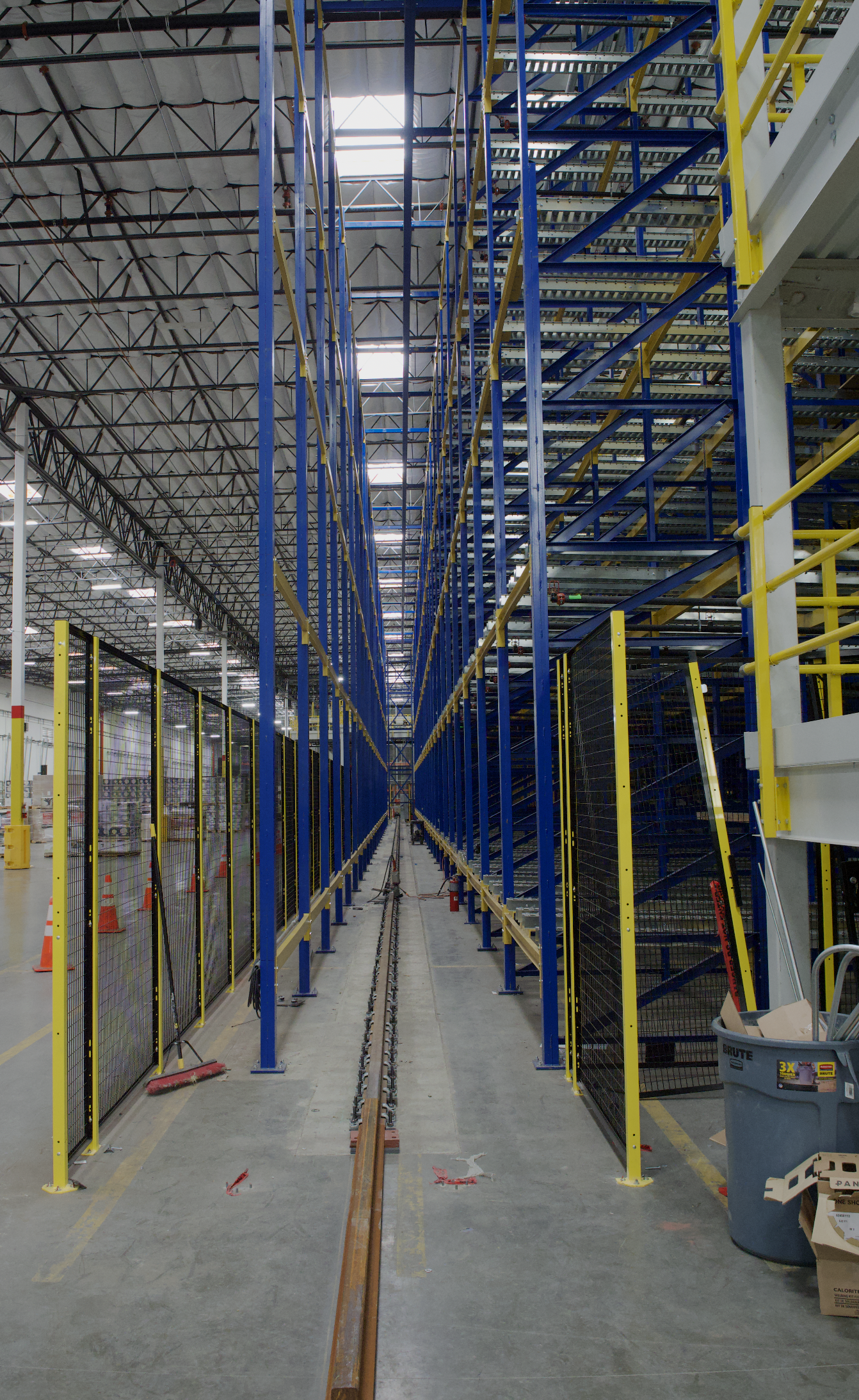 Inside a large warehouse or manufacturing facility with high ceilings, blue and yellow storage racks, and a track running down the center. There are construction tools and orange cones on the floor, and a trash bin with boxes nearby.