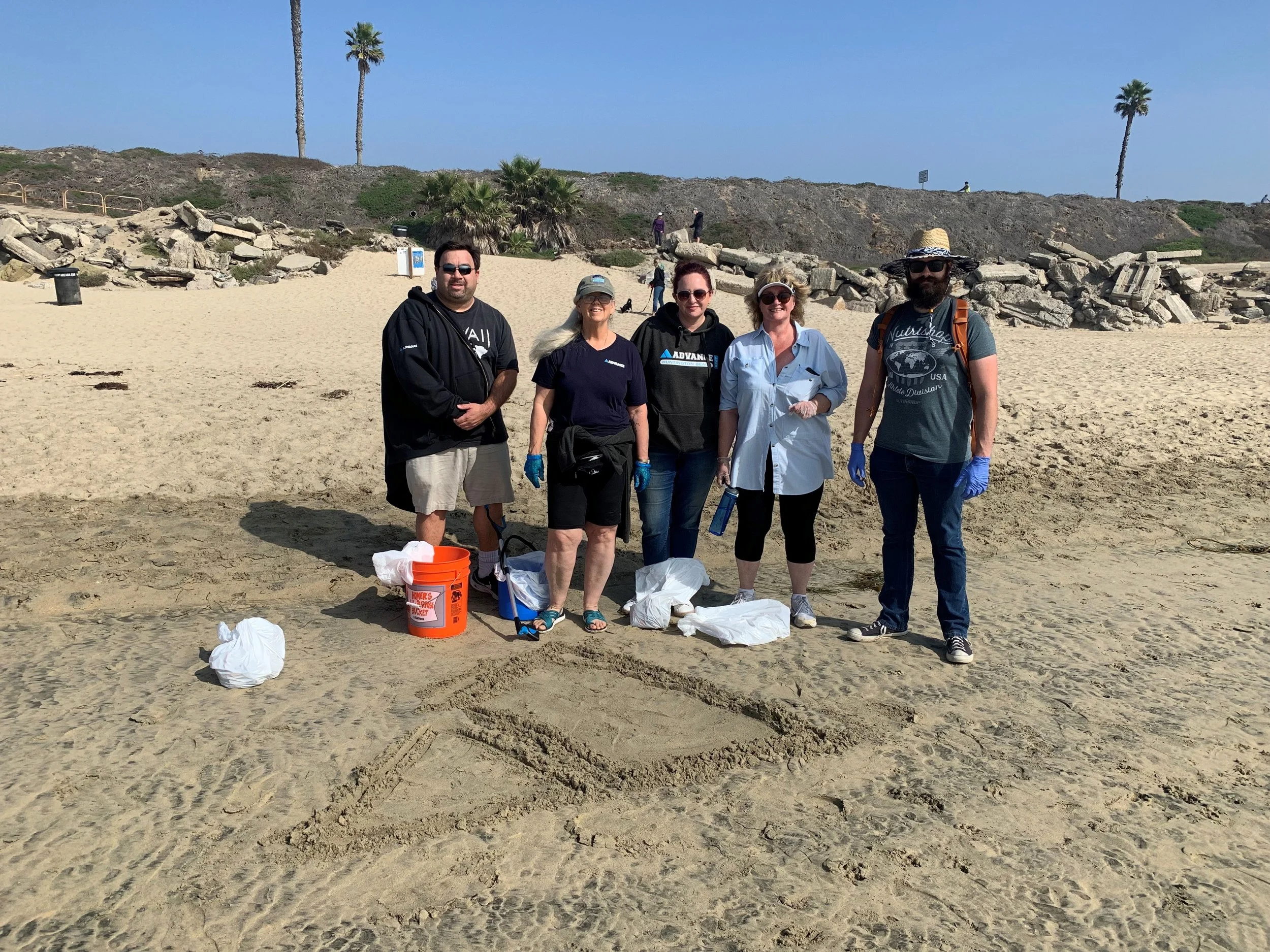 A group of five people standing on the beach with trash bags and supplies, participating in a cleanup activity. There are palm trees and rocky formations in the background, under a clear blue sky.
