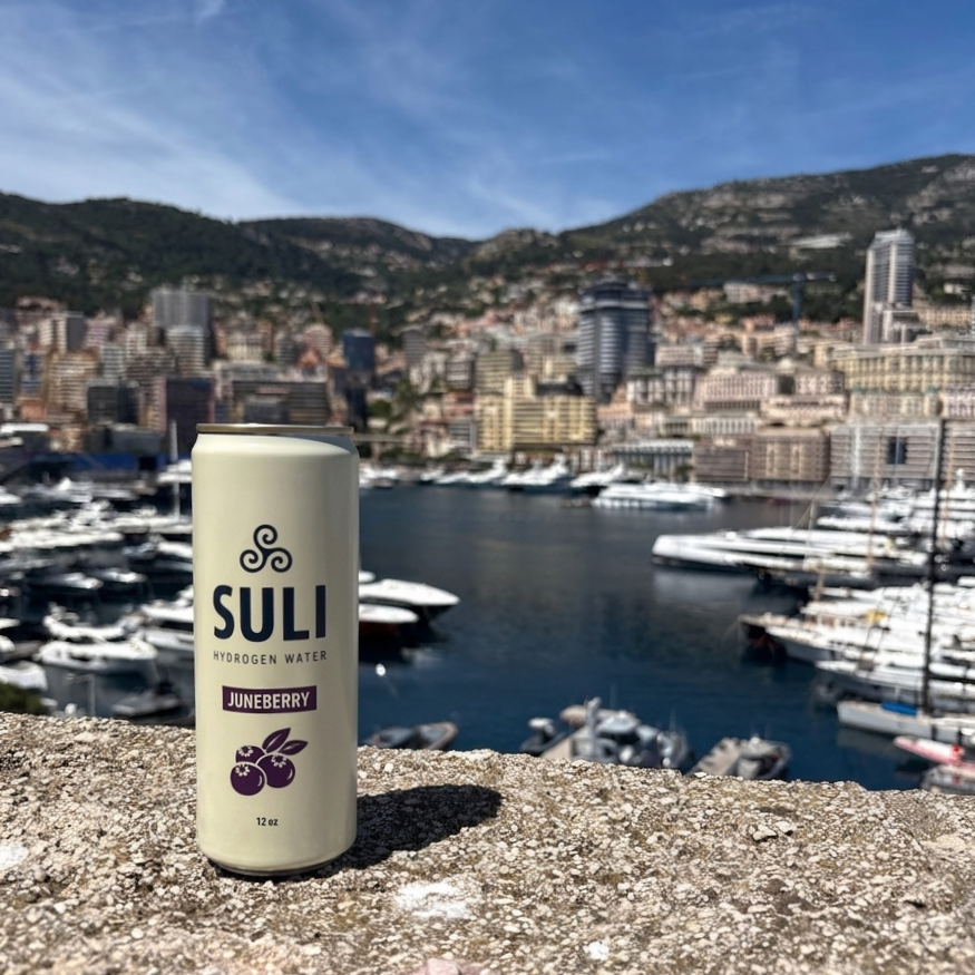 A can of Suli hydrogen water with juneberry flavor on a stone ledge overlooking a marina with yachts, surrounded by city buildings and hills under a blue sky.