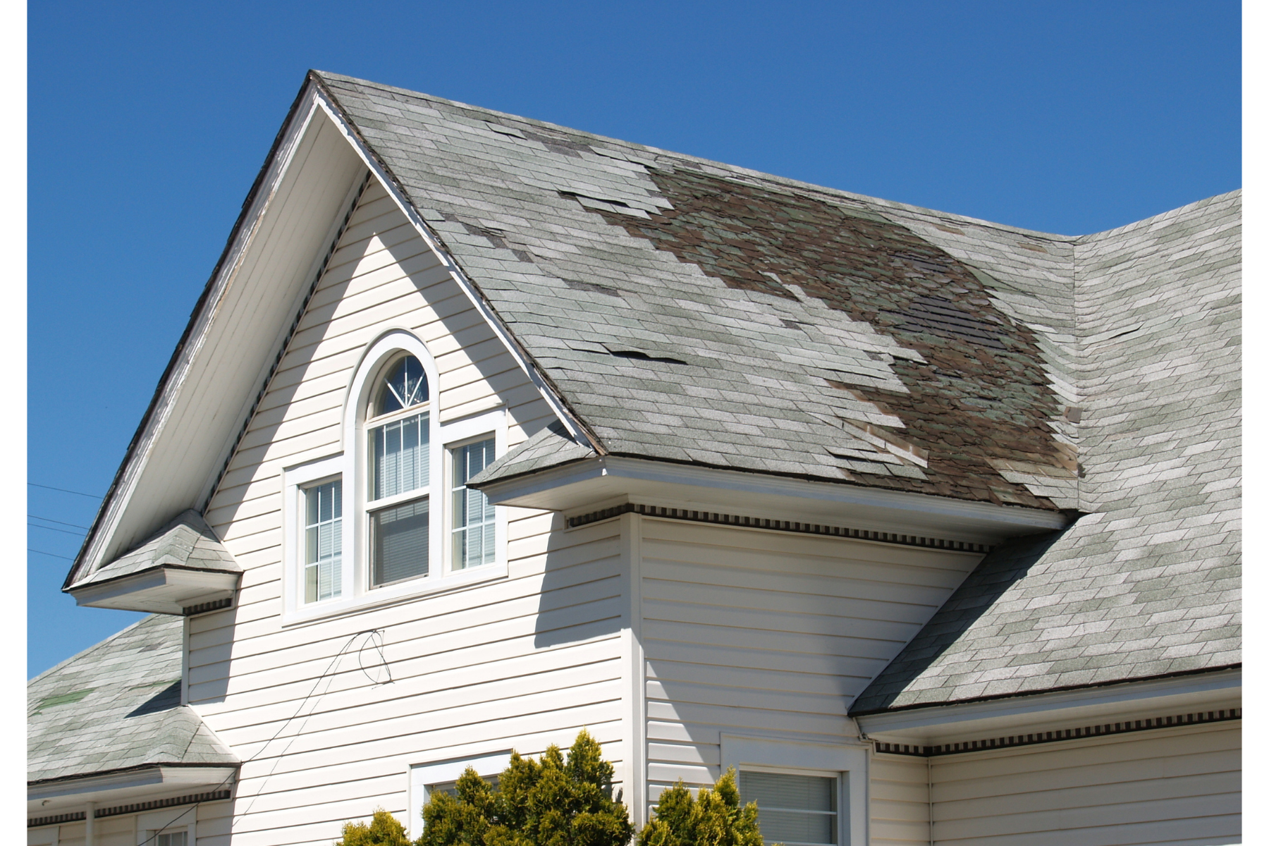 Close-up of a house's roof with damaged and missing shingles, and a white siding wall with a window underneath.