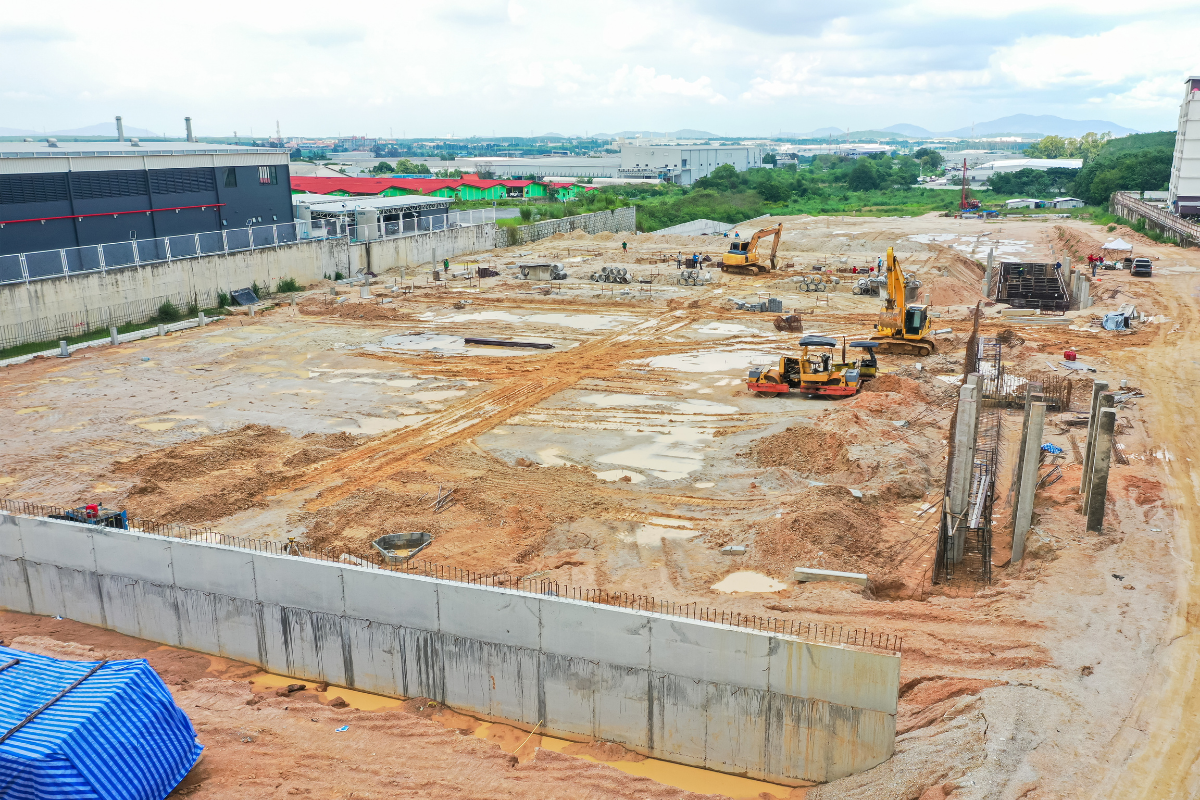 Construction site with heavy machinery, dirt, and building foundations under development, surrounded by industrial buildings and green landscape under a cloudy sky.
