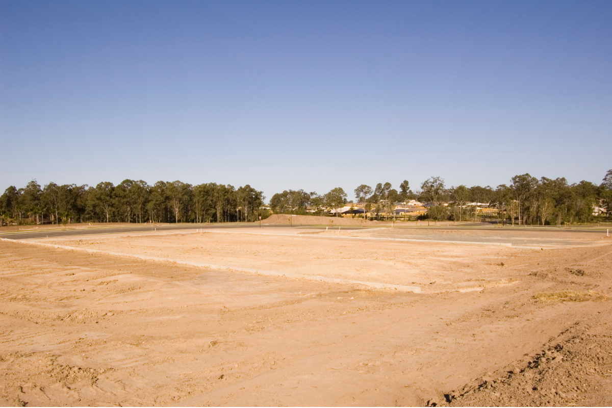 Empty construction site with cleared dirt area, a curved road in the background, and trees and houses beyond.