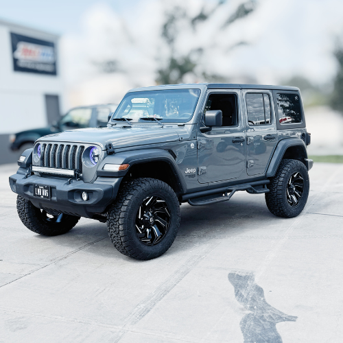 Gray Jeep Wrangler with black wheels parked in dealership lot.