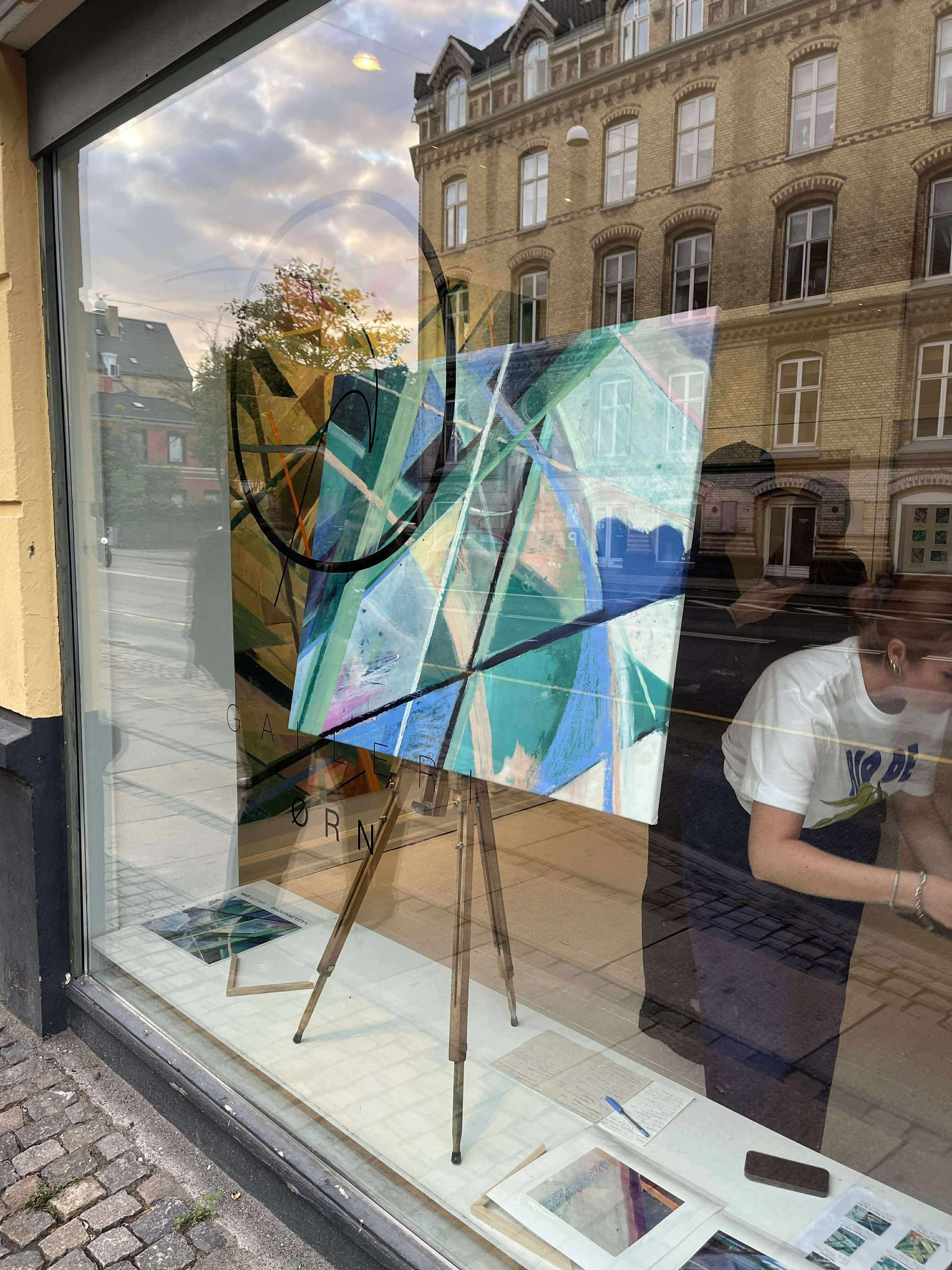 An abstract geometric painting on display in a gallery window, with colorful shapes and lines. The reflection of the street and an adjacent building can also be seen on the glass.