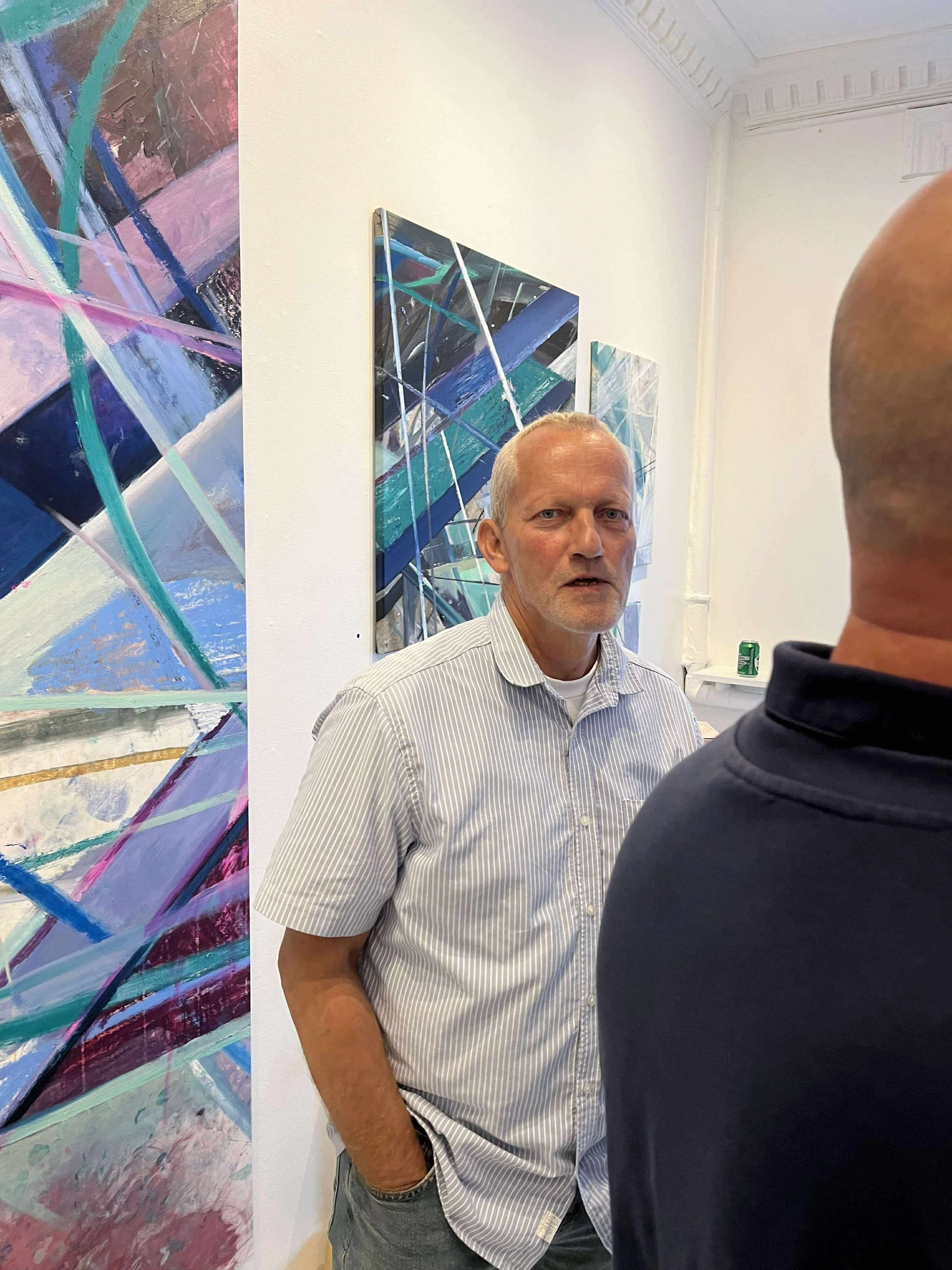 A man in a striped shirt standing and talking in an art gallery with abstract paintings on the wall.